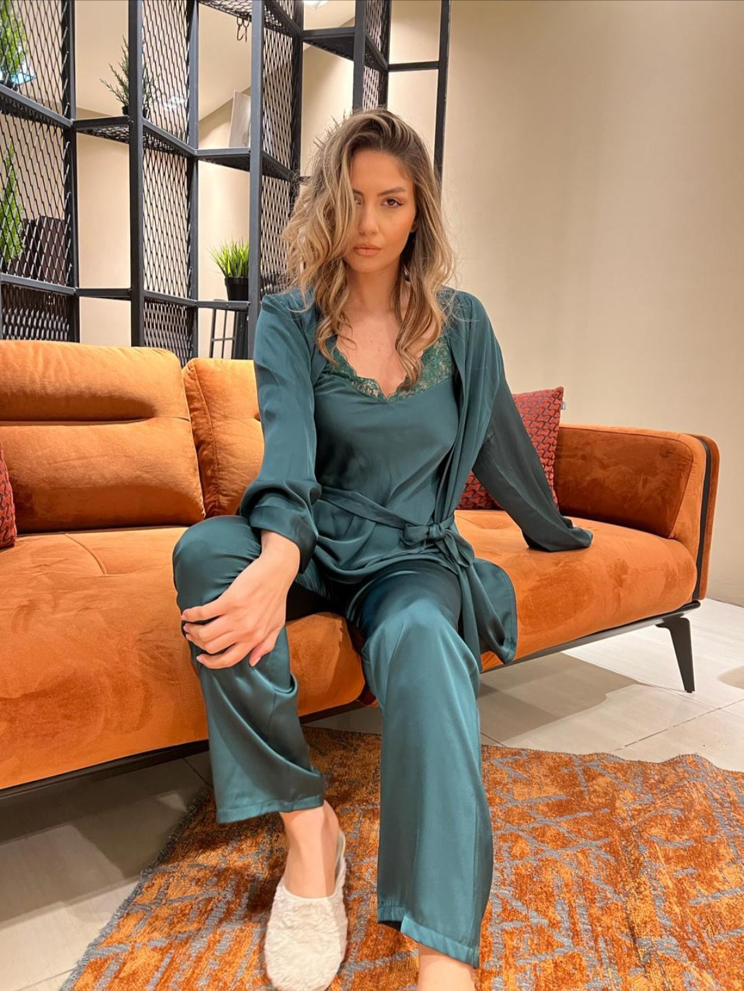 Woman wearing dark olive satin three-piece pants set pyjama, sitting comfortably on an orange couch, enjoying a spring night.