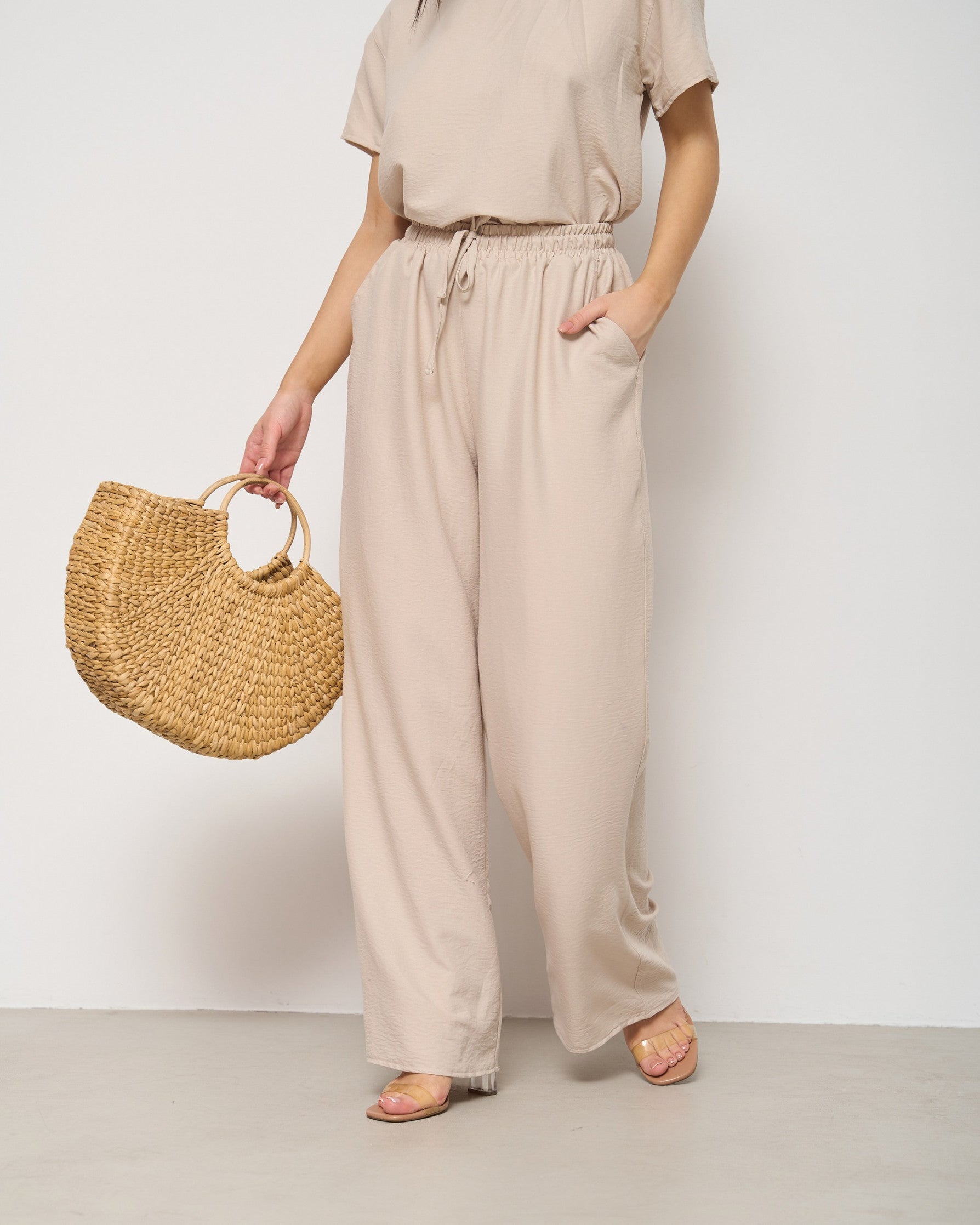 Model wearing beige pyjama set, holding a woven straw bag.