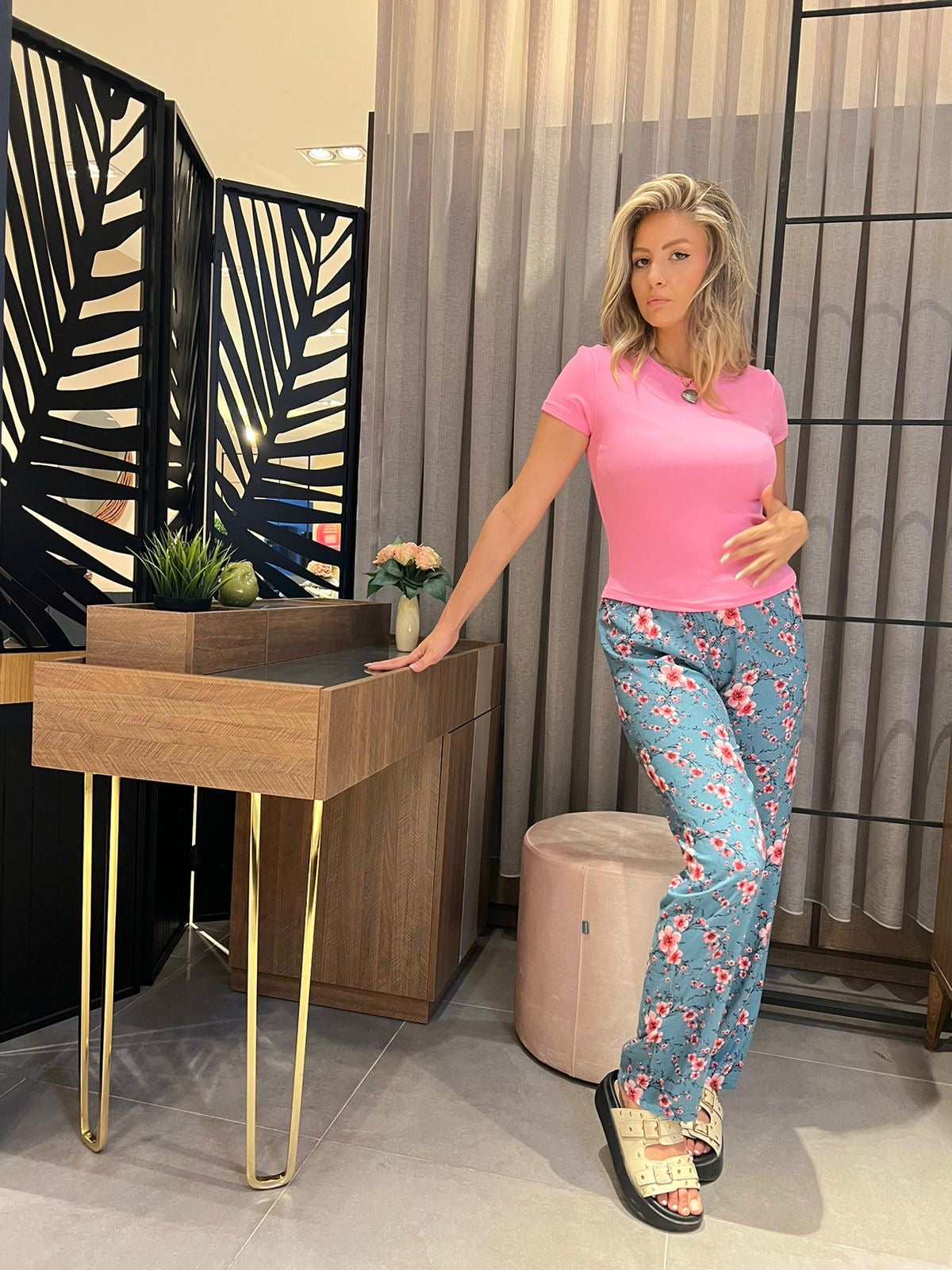 Model wearing a Popjammies pyjama set, featuring a vibrant pink short-sleeve top and floral blue pants, standing stylishly next to a modern wooden vanity with decorative elements.