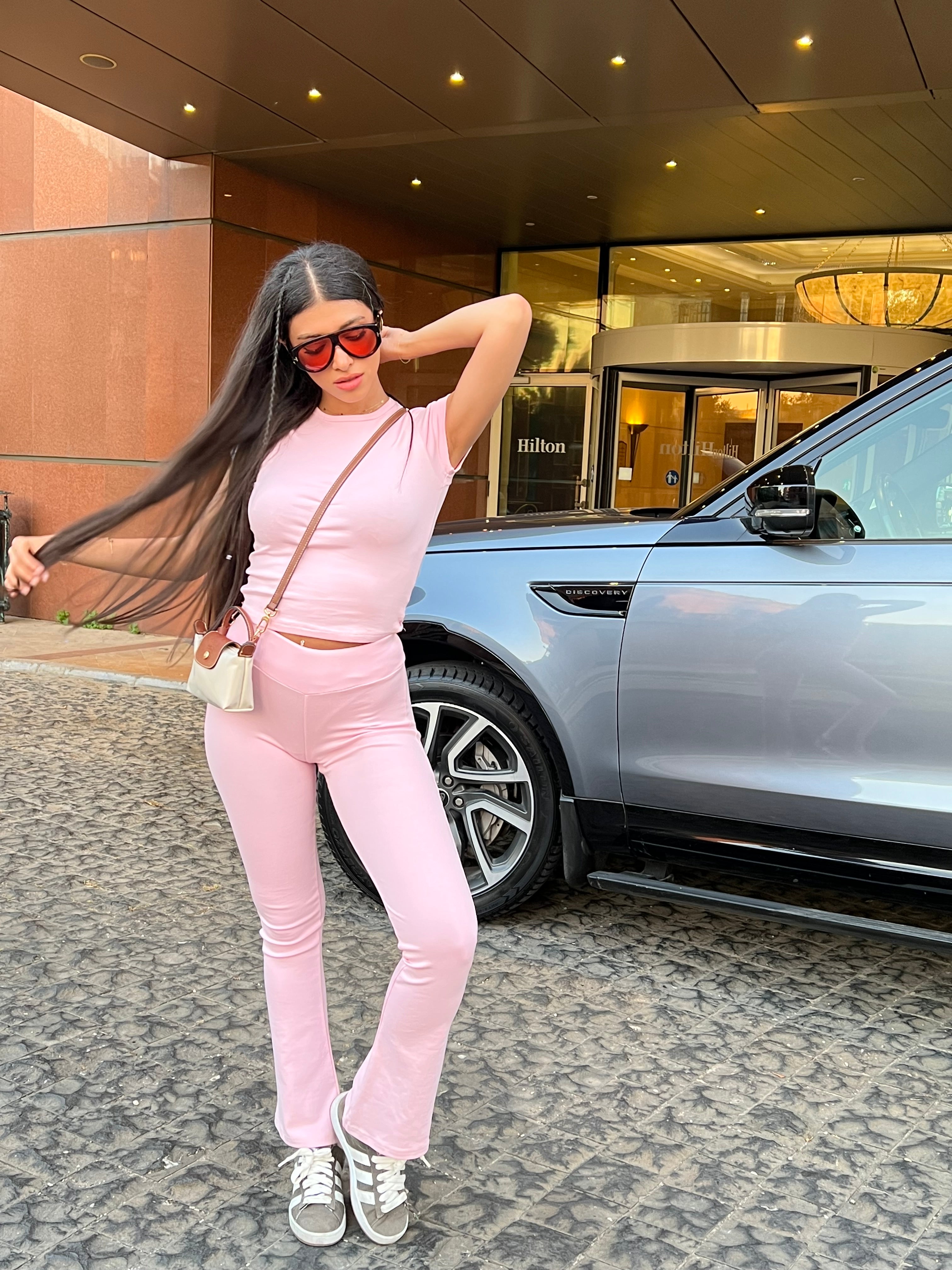 Woman in a pink pajama set with a fitted T-shirt and flared pants, standing next to a grey car in front of the Hilton hotel.