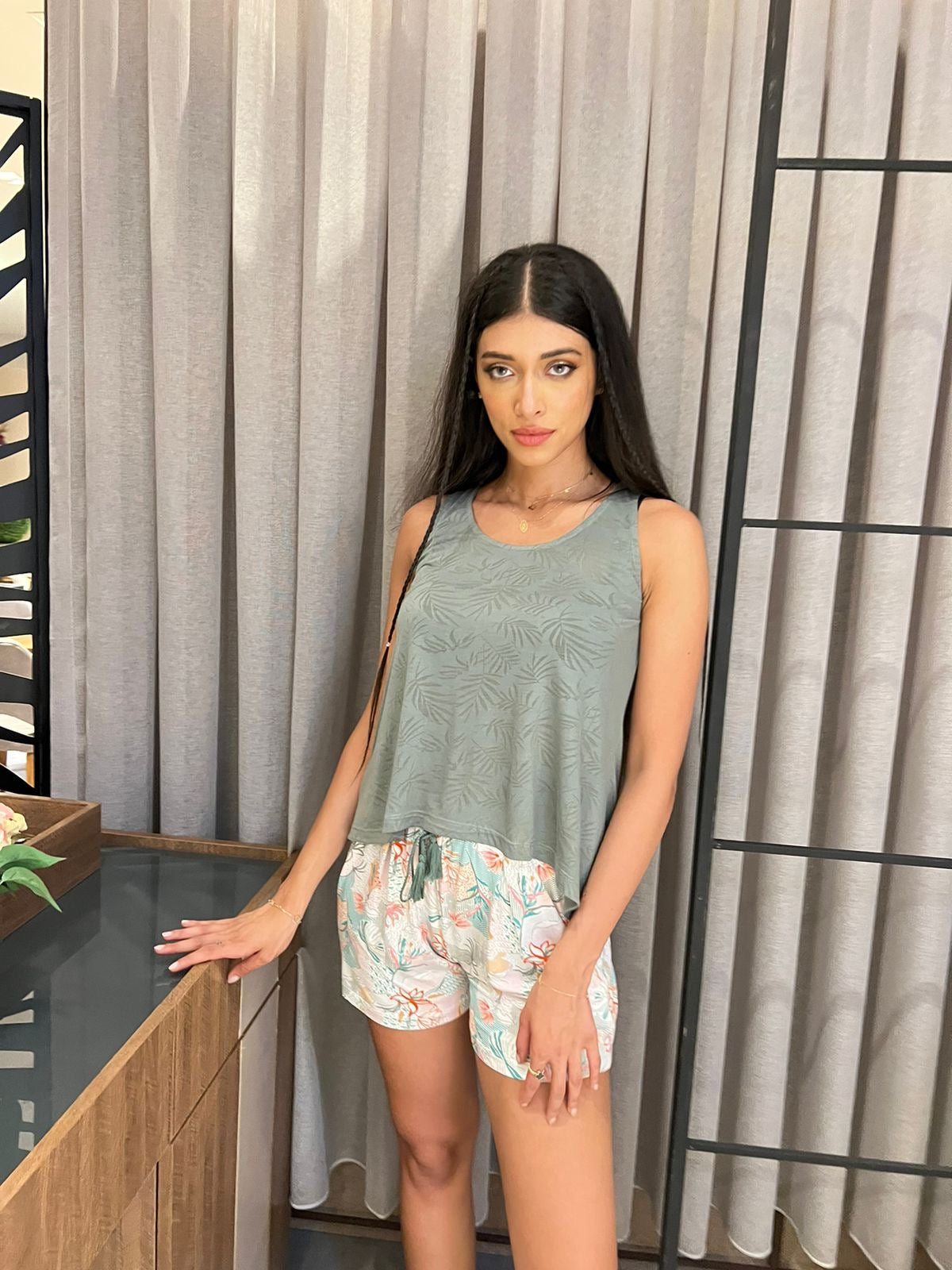 Model in a green tank top and floral print shorts pajama set, standing against a modern curtain backdrop.