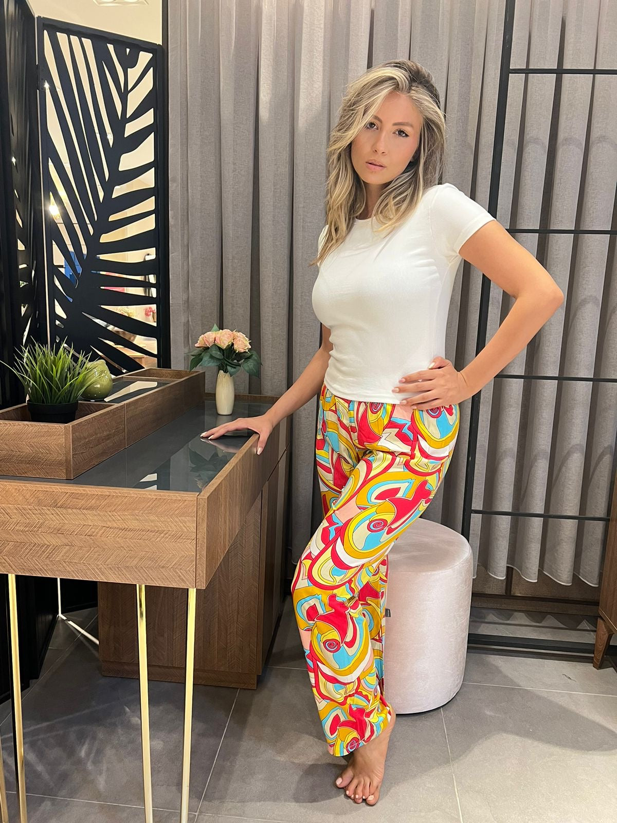 Model wearing a stylish Popjammies pyjama set, featuring a crisp white short-sleeve top paired with vibrant, multicolored patterned pants, standing next to a modern wooden vanity with decorative elements.