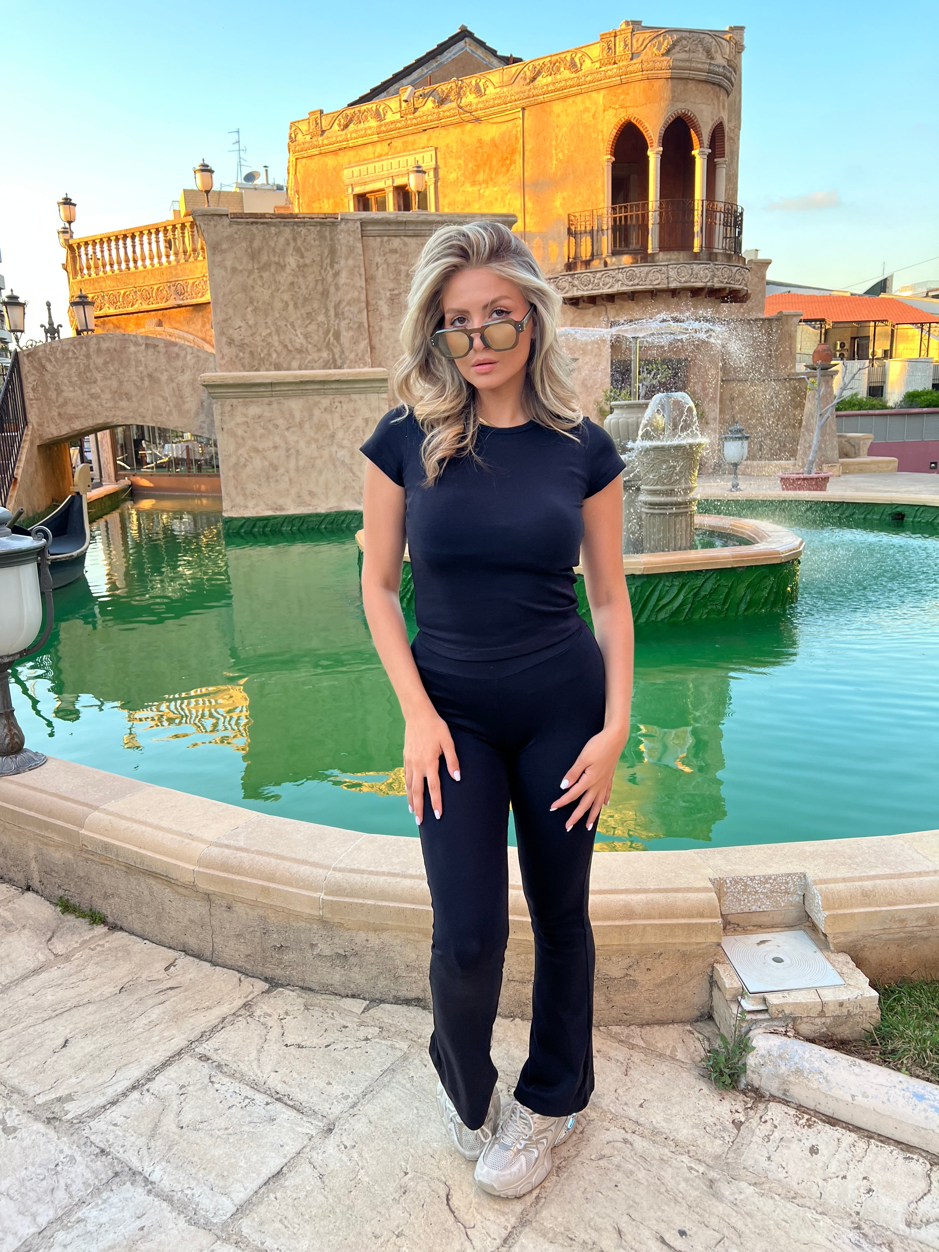 Woman in a black pajama set with a fitted T-shirt and flared pants, standing in front of a green fountain with a historical building in the background.