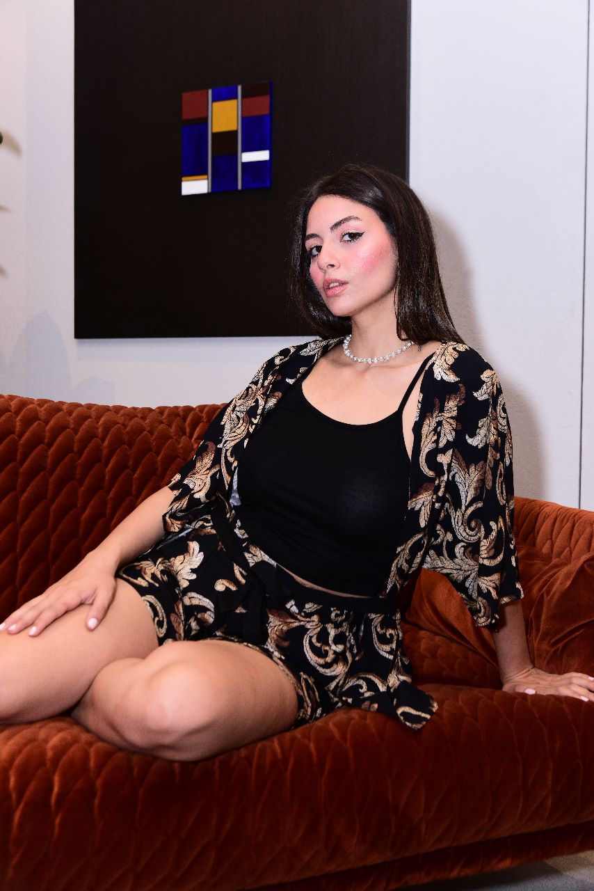 Model sitting on an orange velvet sofa, wearing a black and gold patterned pyjama set by Popjammies, featuring a black camisole and matching shorts with a kimono-style robe.