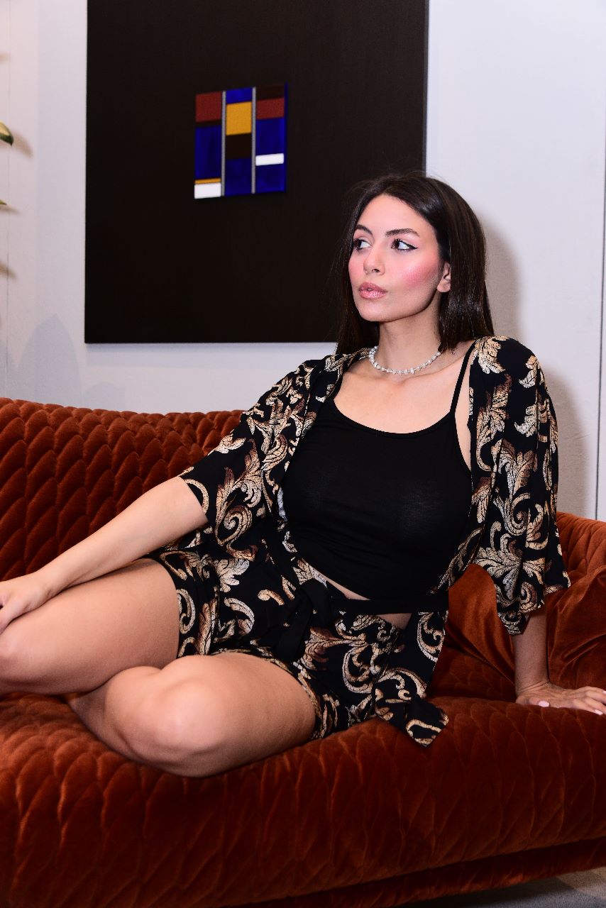 Model sitting on an orange couch, showcasing a stylish black and gold paisley pyjama set by Popjammies. The set includes a loose kimono-style robe and matching shorts, paired with a black tank top and accessorized with a pearl necklace.