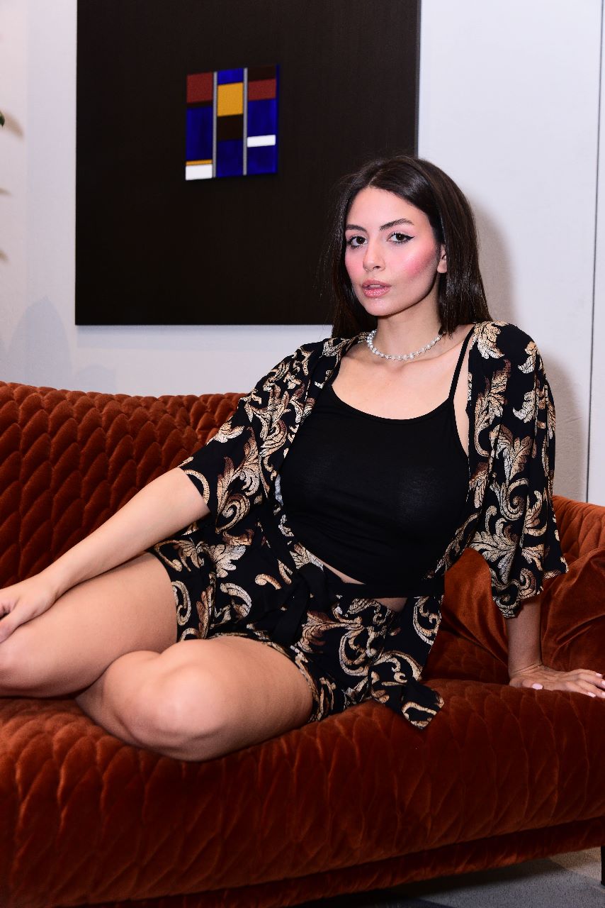 Model sitting on an orange couch, wearing a stylish black and gold paisley pyjama set by Popjammies, featuring a loose kimono-style robe and matching shorts, complemented by a black top and a pearl necklace.