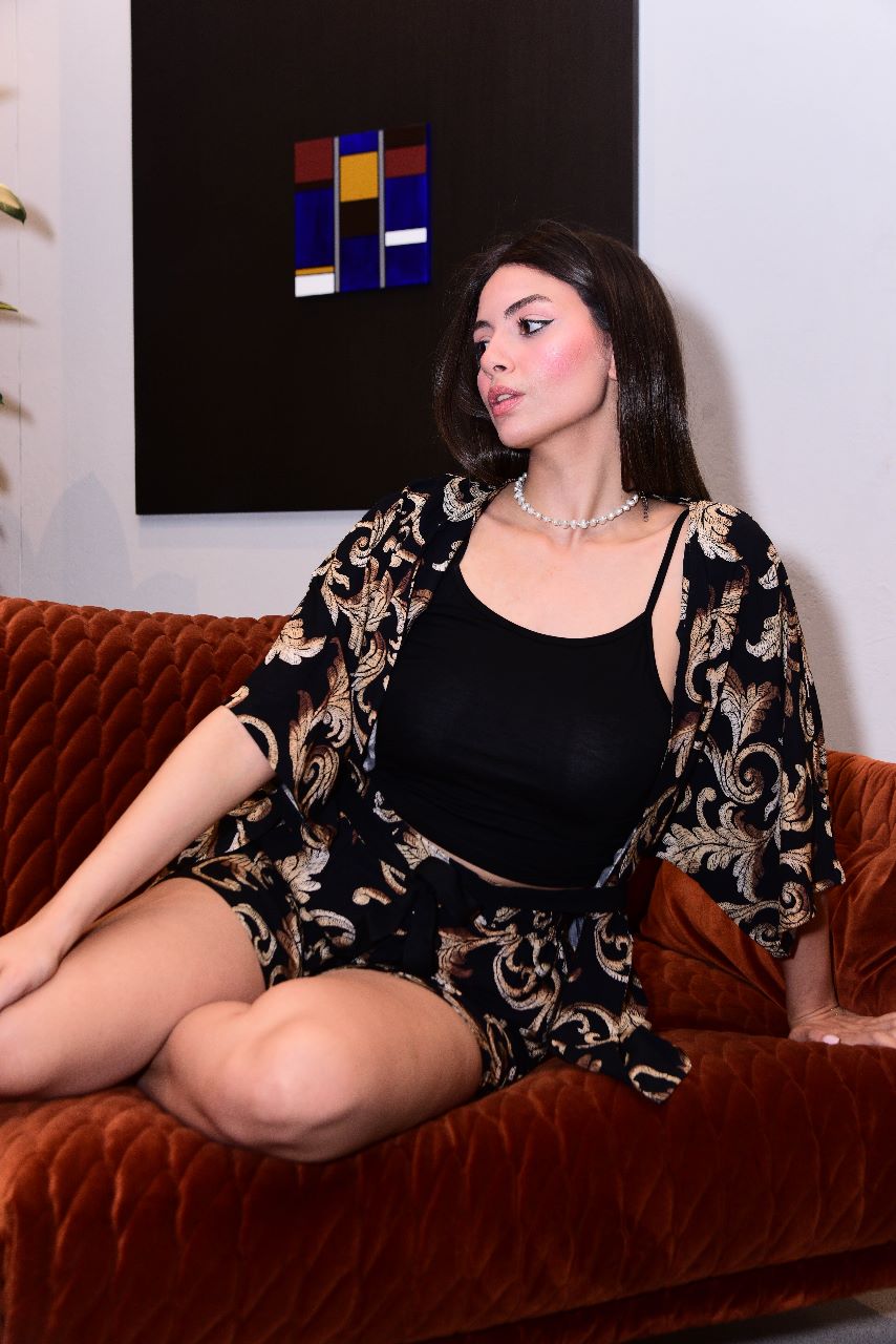 Model lounging on a plush orange couch, wearing a luxurious black and gold paisley pyjama set by Popjammies, consisting of a flowy kimono-style robe and matching shorts, paired with a black top.