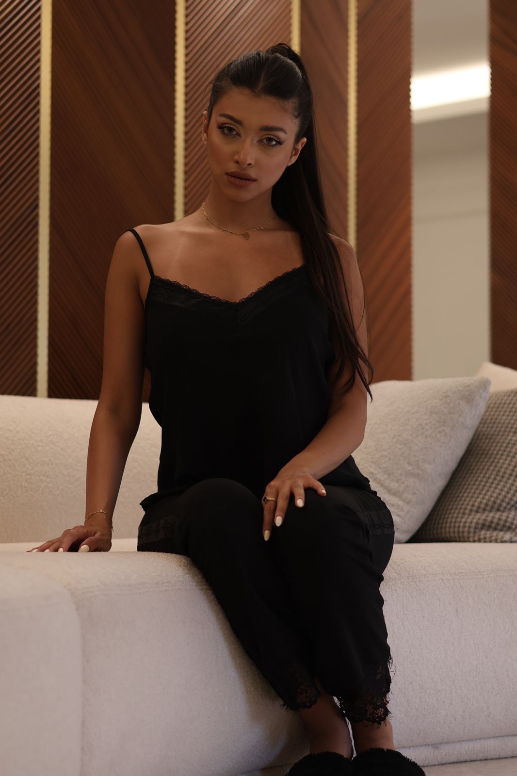 Woman seated on a light-colored sofa, wearing a black camisole and pajama set with lace trim, looking directly at the camera in a cozy indoor setting.