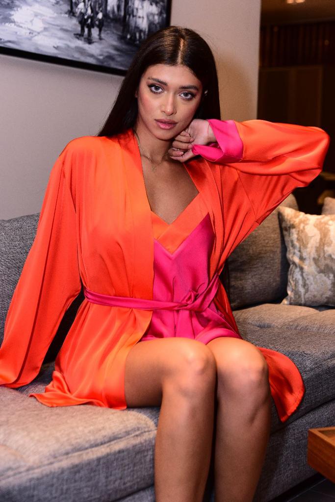 Model sitting on a sofa, wearing a vibrant satin pyjama set by Popjammies, featuring a hot pink camisole and an orange robe, looking stylish and comfortable.