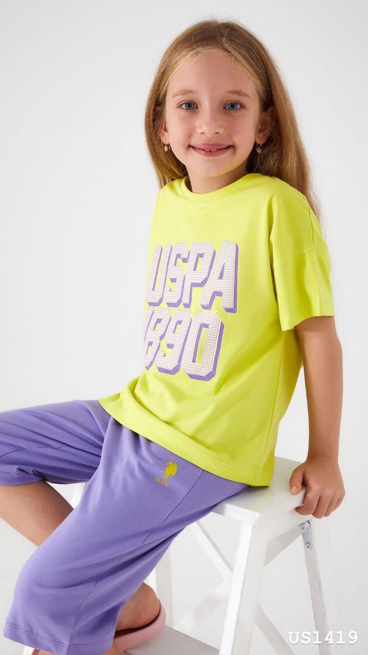 Girl sitting on a white stool, wearing a yellow and purple pyjama set with 'USPA 1890' print from Popjammies