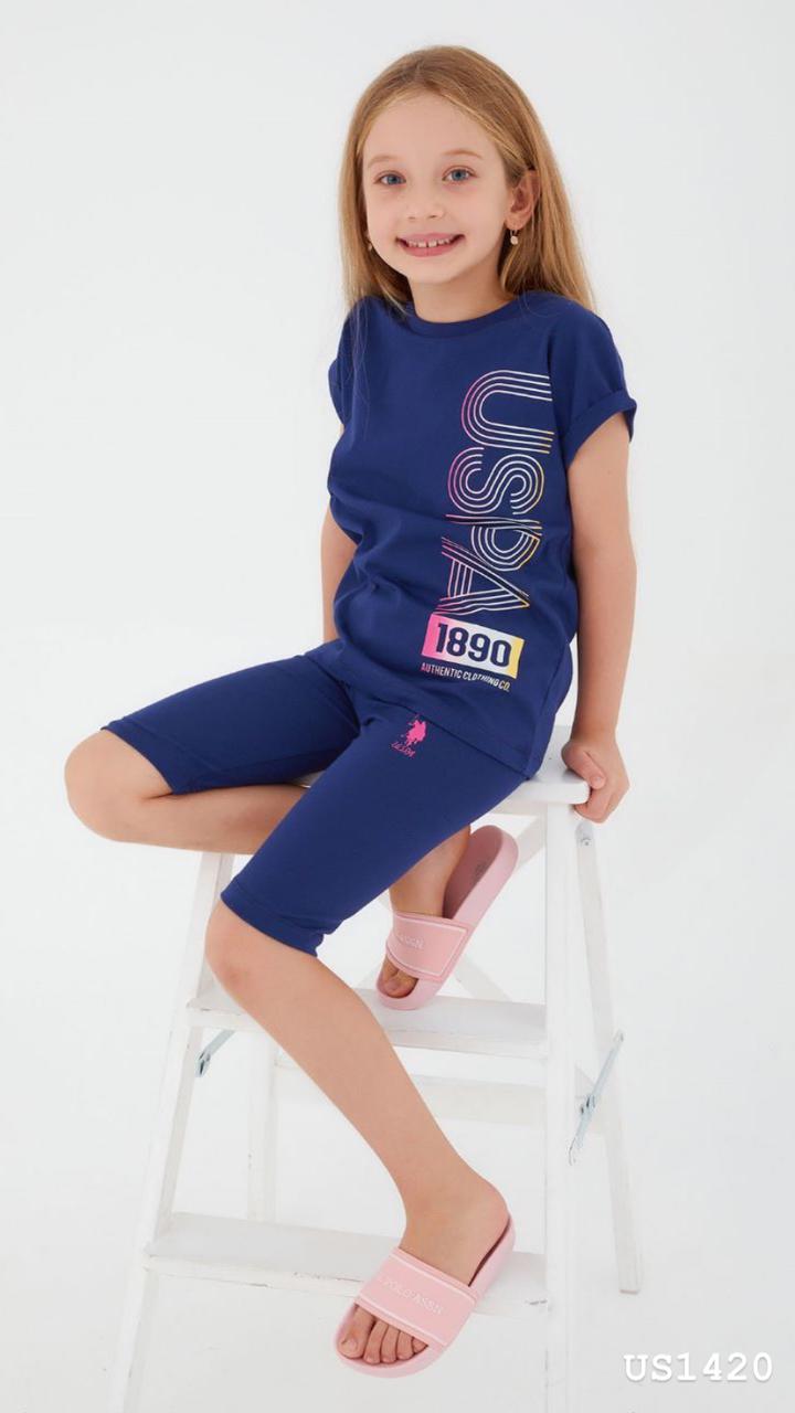 Child wearing the Popjammies Blue Horizon Pajama Set, featuring a vibrant blue T-shirt with a "USPA 1890" vertical graphic and matching blue shorts, smiling and sitting on a white chair.