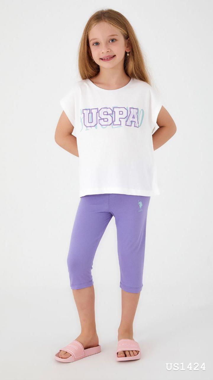 Child wearing the Popjammies Lilac Dream Pajama Set, featuring a white T-shirt with a "USPA" graphic and lilac capri pants, smiling and looking comfortable.