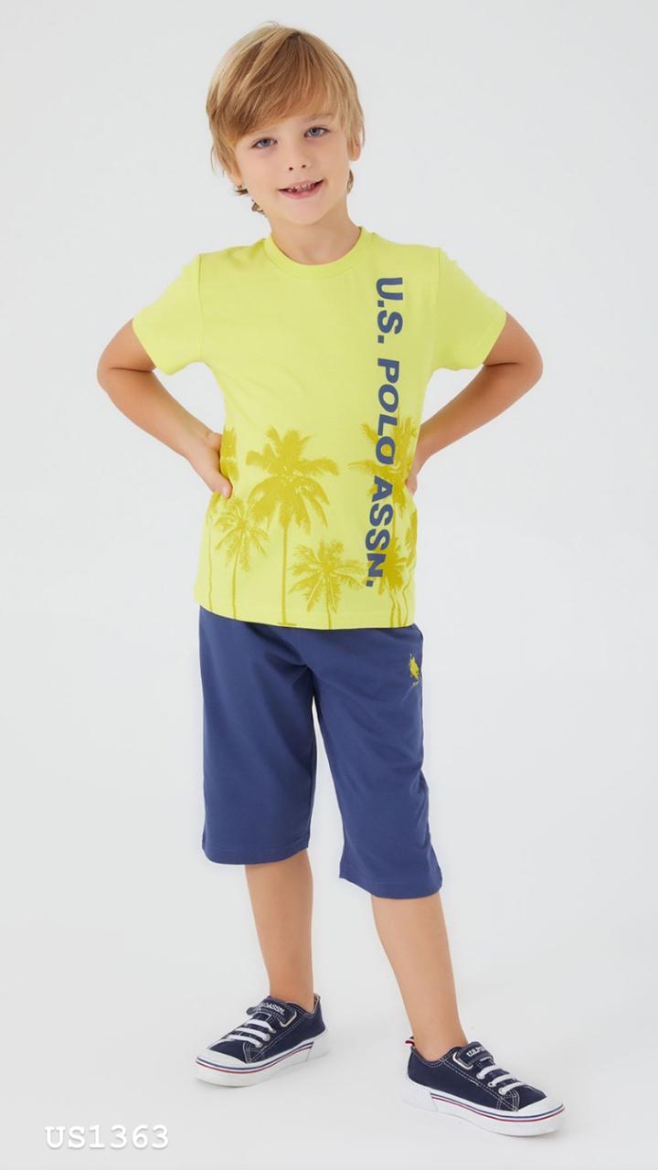 Smiling boy wearing a yellow top with "U.S. POLO ASSN." logo and palm tree graphic, paired with navy blue shorts pyjama set from Popjammies.