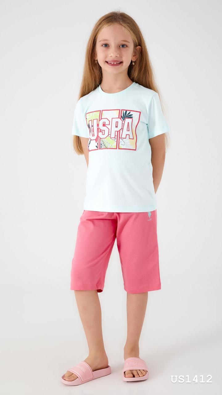 Smiling girl wearing a light blue pyjama top with 'USPA' logo and tropical motif, paired with bright pink shorts.