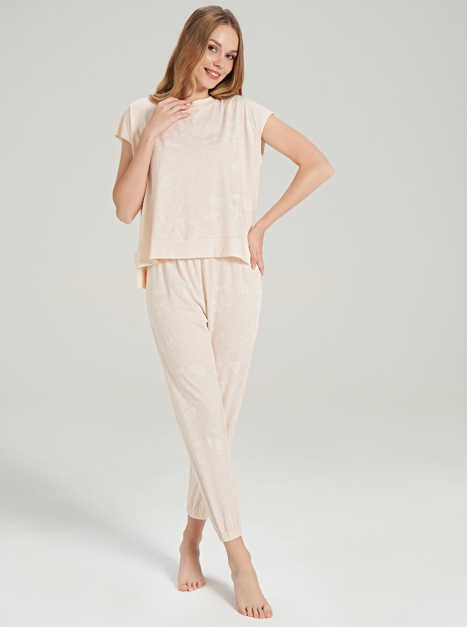 A woman standing and modeling a beige floral pajama set with a relaxed fit top and matching pants.