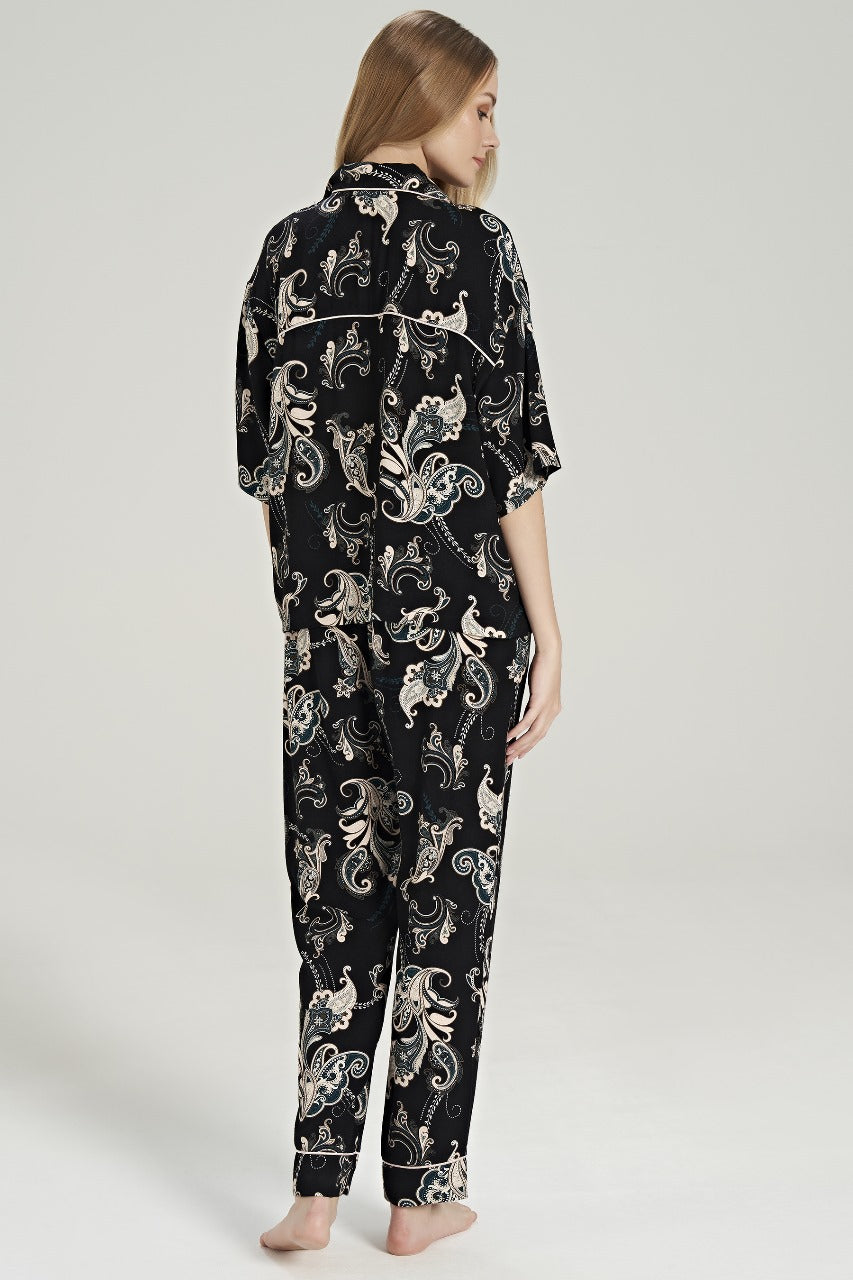 Rear view of a model wearing the Popjammies Women's Black Paisley Print Pyjama Set, featuring a soft, button-up top with a notched collar and short sleeves, and matching relaxed-fit pants adorned with an intricate paisley pattern in shades of cream and teal against a black background.