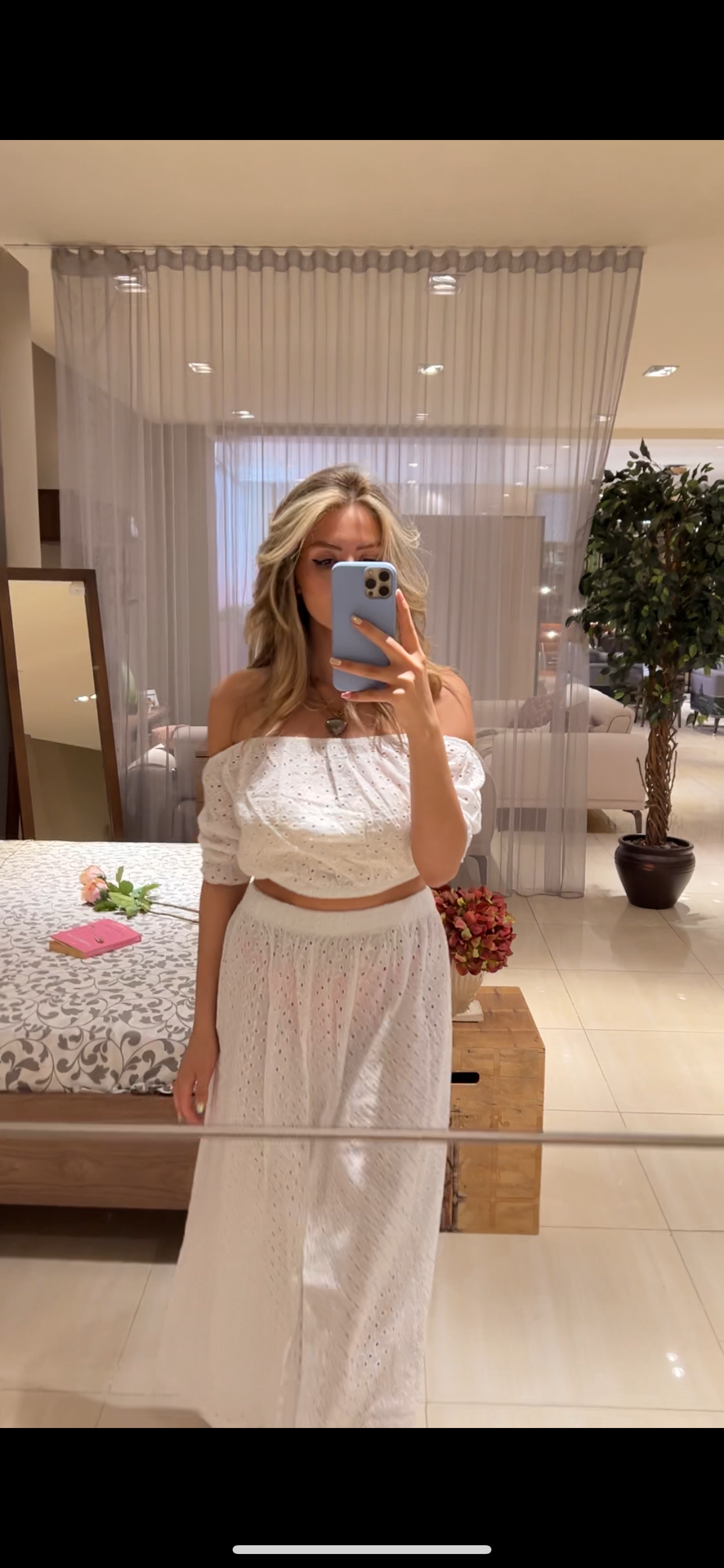 Woman wearing Popjammies Women's Eyelet Off-Shoulder White Pyjama Set, taking a mirror selfie in a cozy room. The set includes an off-shoulder top with eyelet details and a matching full-length skirt, both in white.