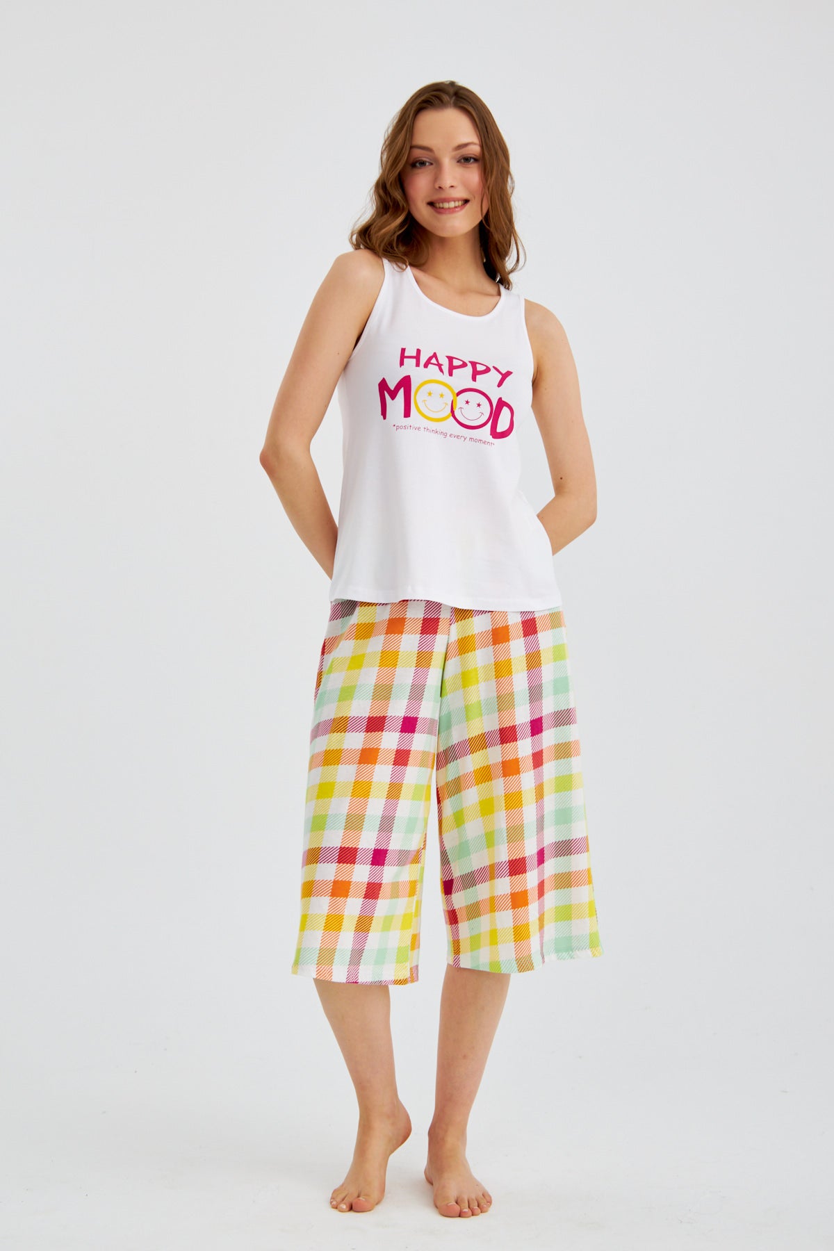 Woman wearing Popjammies "Happy Mood" Pyjama Set with a white sleeveless top and multicolored plaid wide-leg pants, standing and smiling.