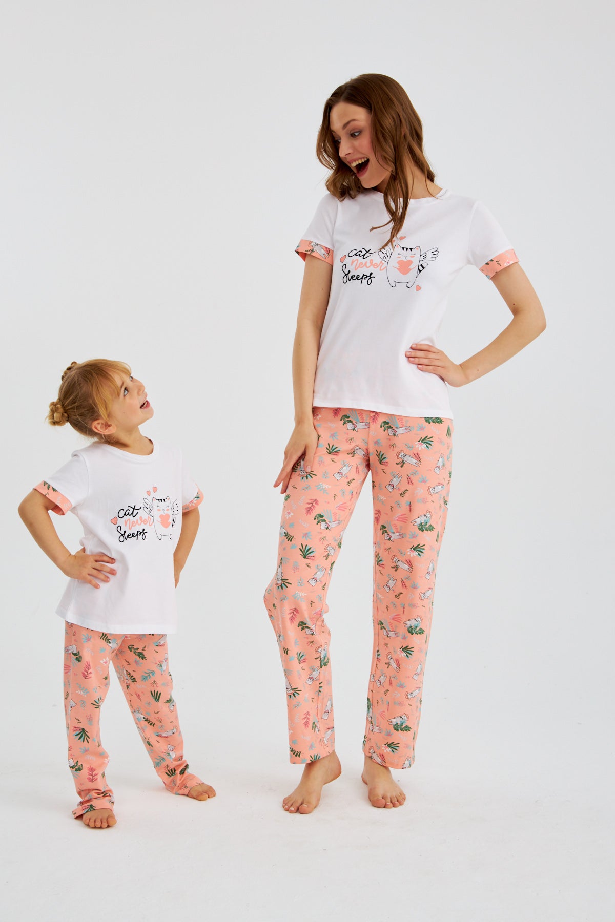 Mother and daughter wearing matching Popjammies "Cat Never Sleeps" pyjama set with white tops and pink floral print pants, smiling and looking at each other.