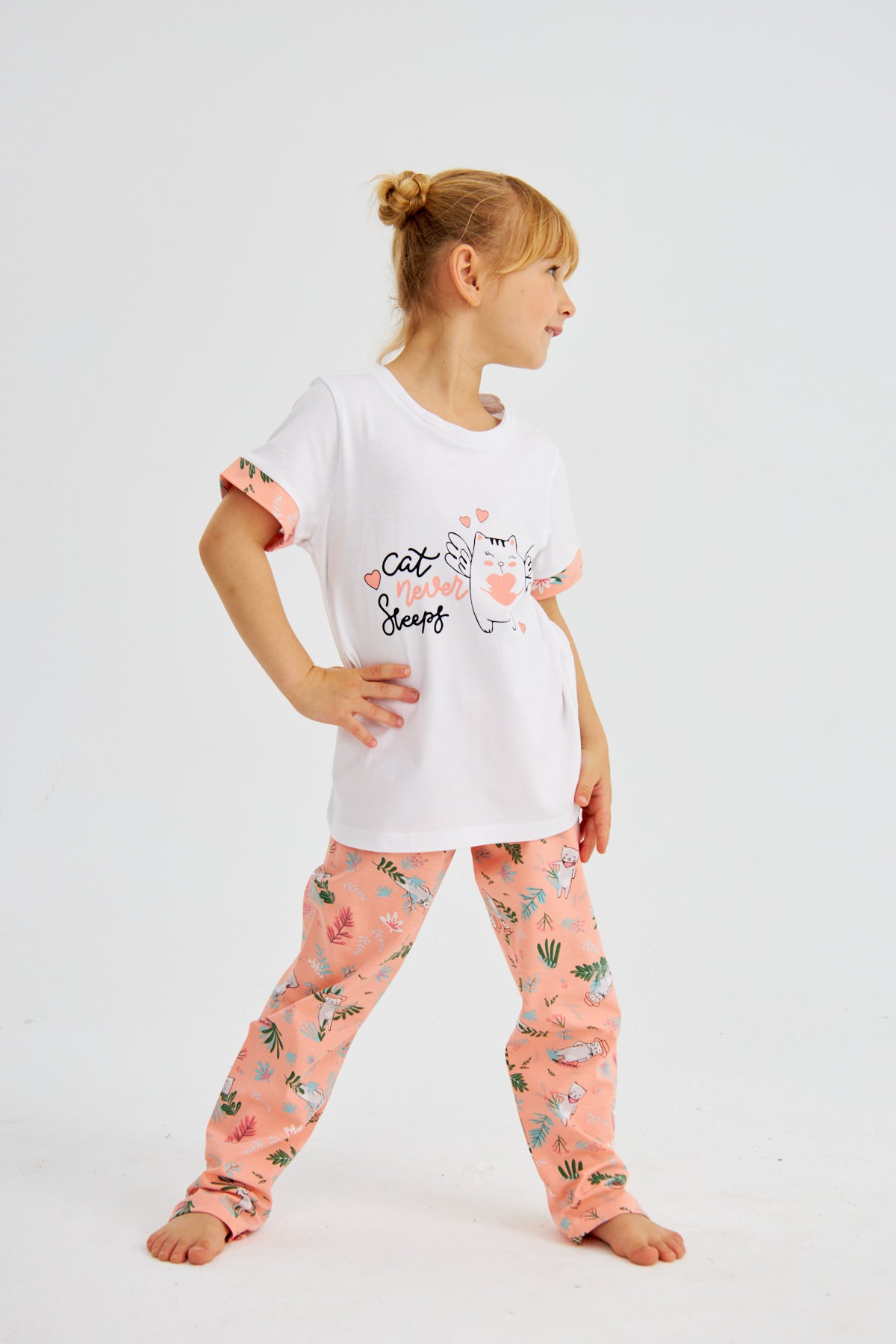 Young girl standing in Popjammies "Cat Never Sleeps" pyjama set, white top with cat graphic and pink floral print pants, looking to the side.