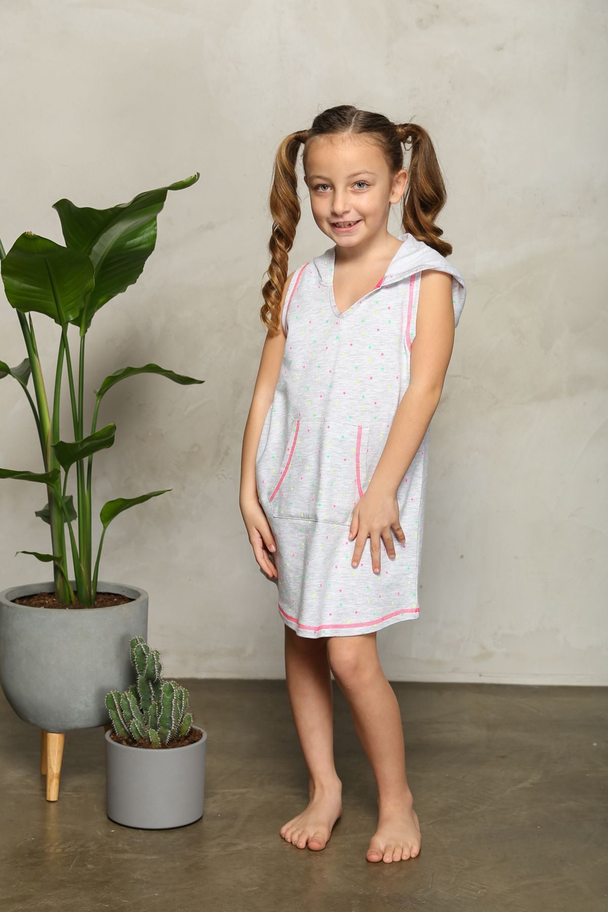 Young girl smiling and posing in sleeveless hoodie dress with pastel polka dots, standing near plants.