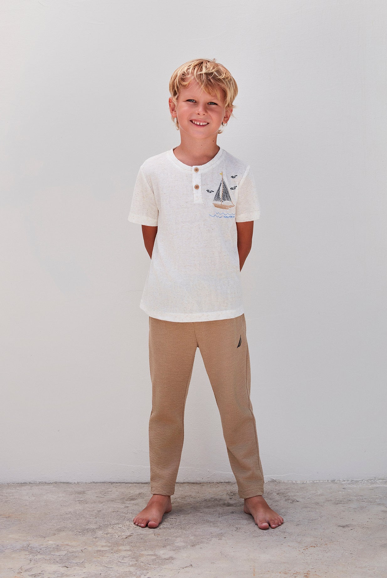  A boy wearing a white top with a sailboat design and beige pants, standing against a white background.