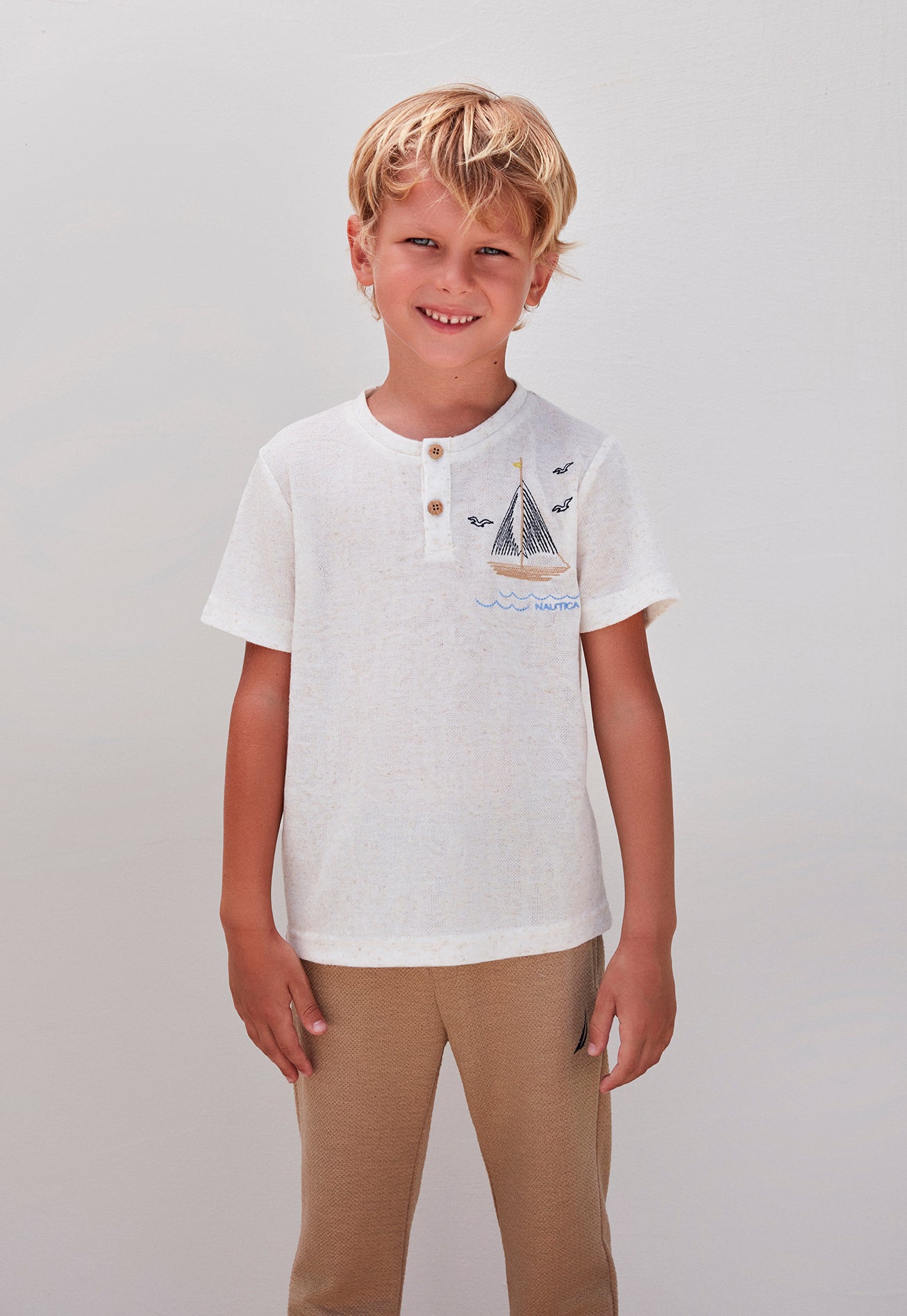 A smiling boy in a white top with a sailboat design and beige pants, posing against a white background.