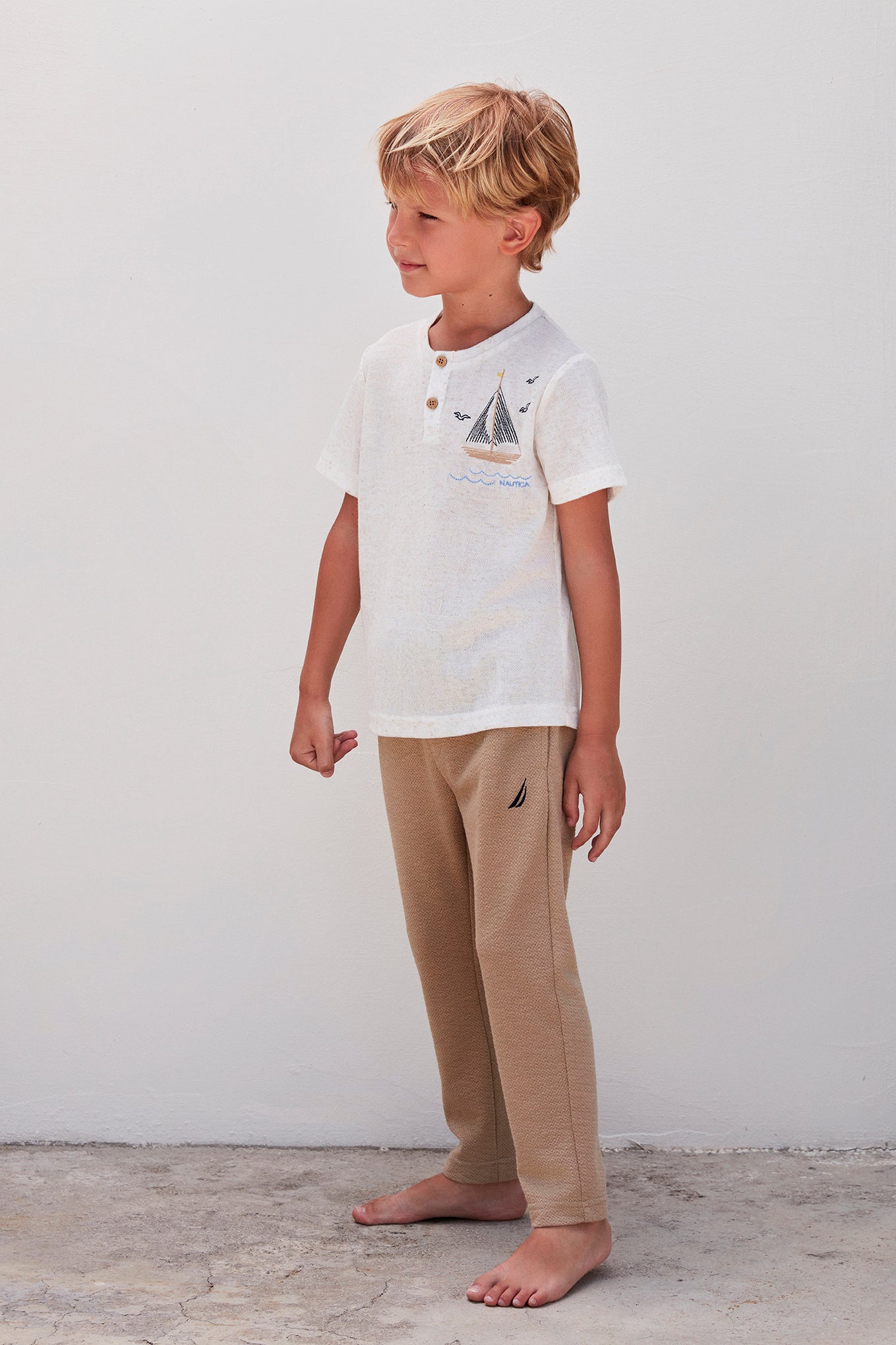 A boy looking to the side, wearing a white top with a sailboat design and beige pants, standing against a white background.