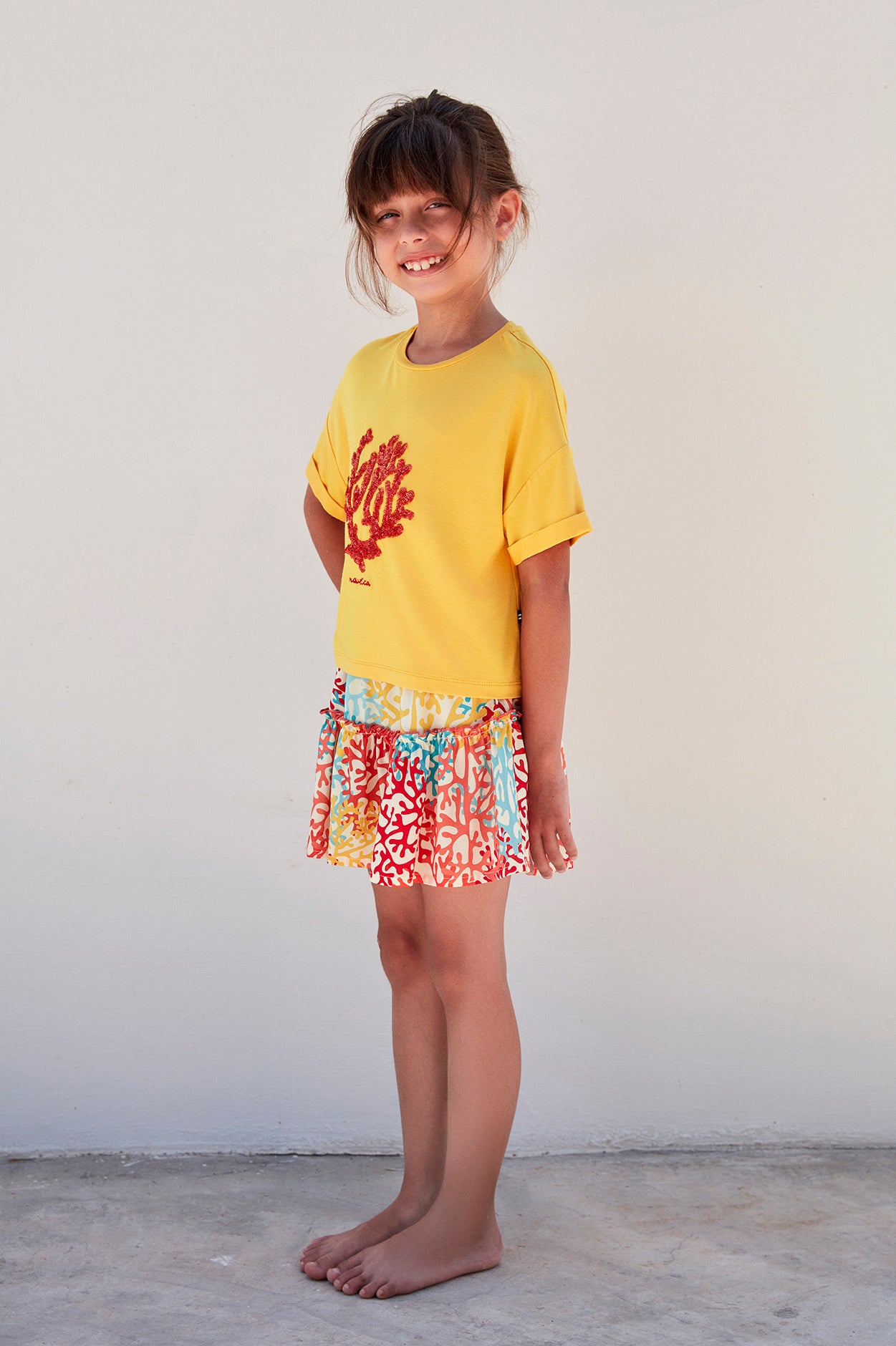 Child wearing a yellow top with a red coral design and a multicolored coral reef print skirt, standing and smiling