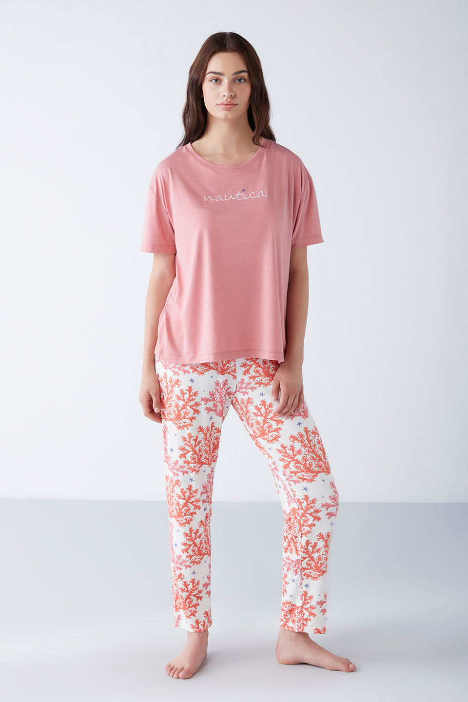 Model wearing Popjammies Women's Pyjama Set with a pink T-shirt and white pants with orange coral reef print, standing and looking straight ahead.