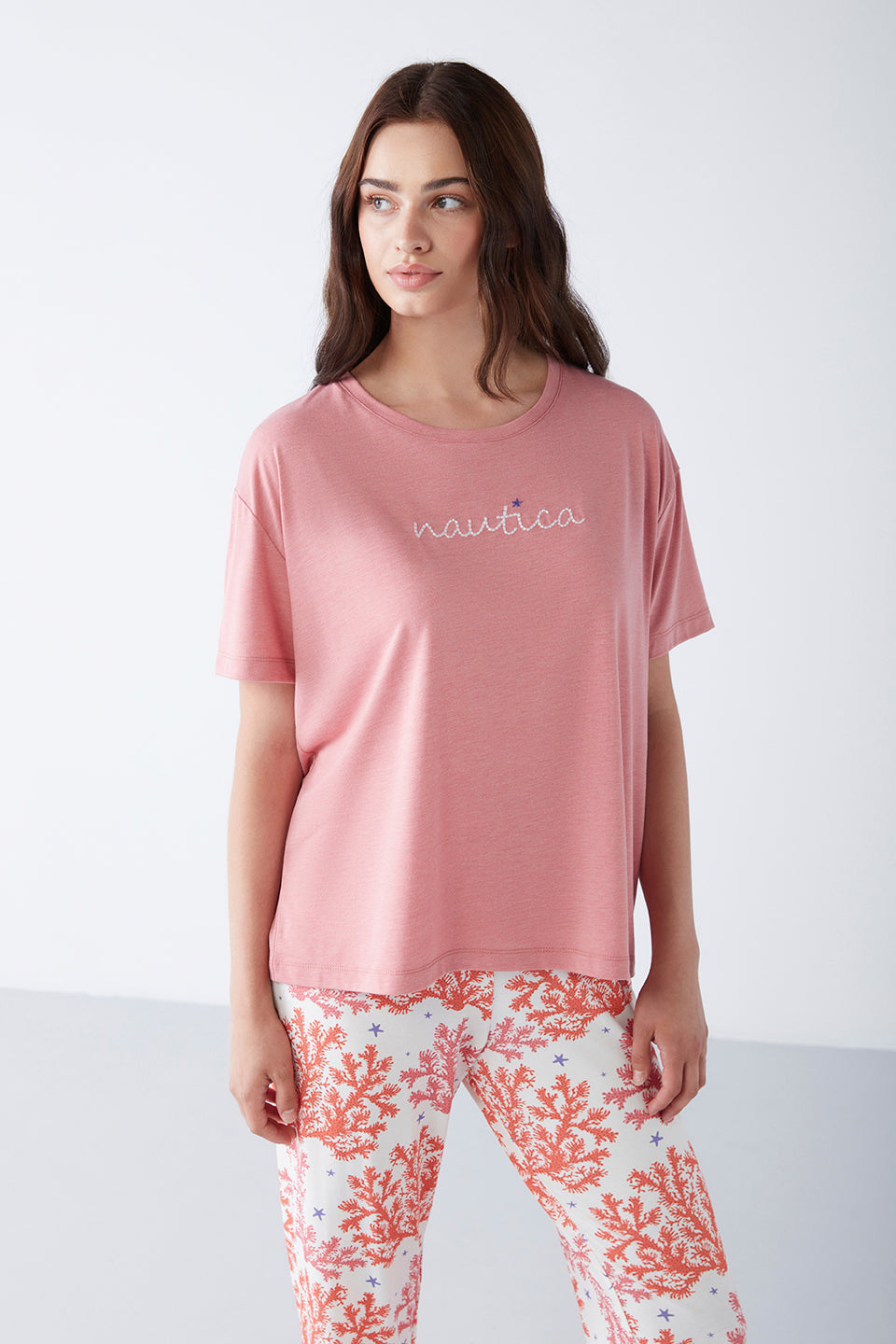 Model wearing a Popjammies Women's Pyjama Set with a pink T-shirt and white pants featuring an orange coral reef print, looking off to the side.