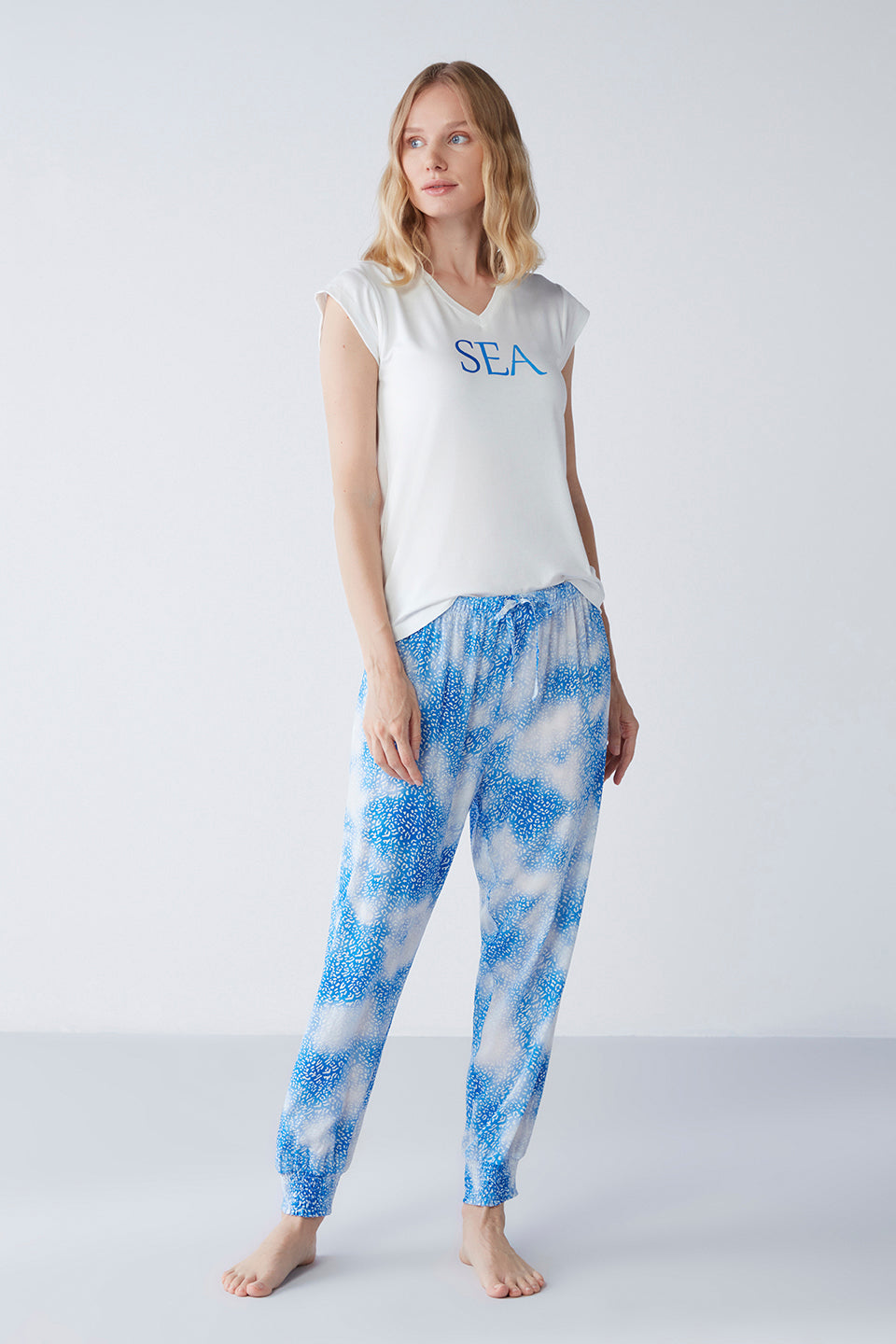 Woman wearing Popjammies "Sea Breeze" Pyjama Set, featuring a white V-neck top with "SEA" print and blue patterned pants, standing in a minimalistic white room.