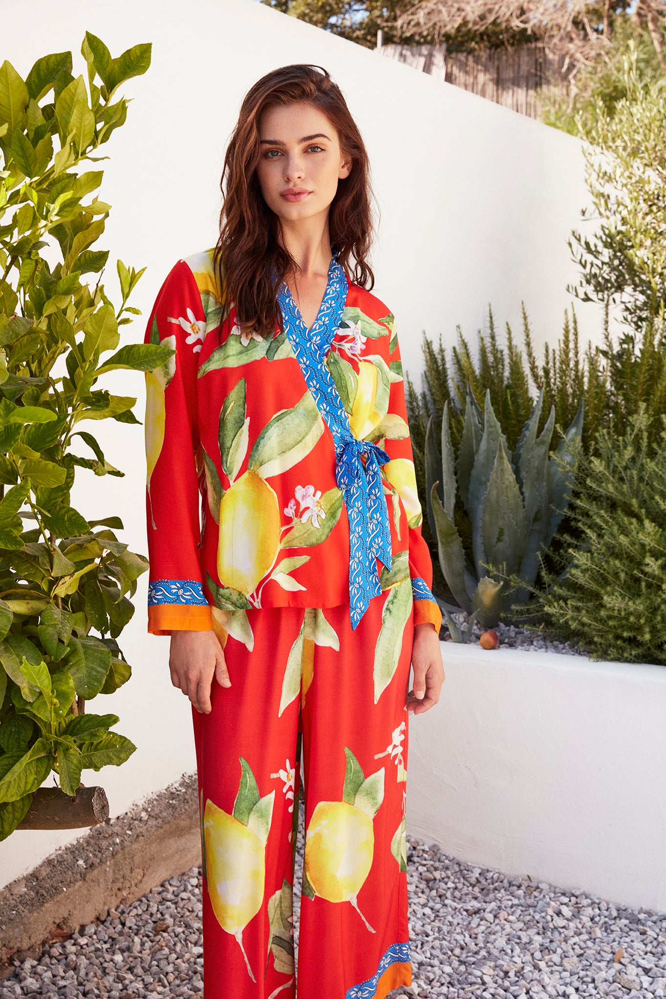 Woman wearing Popjammies' Citrus Bloom Pyjama Set with vibrant lemon prints, a wrap-around top with a blue floral tie, and wide-leg pants.