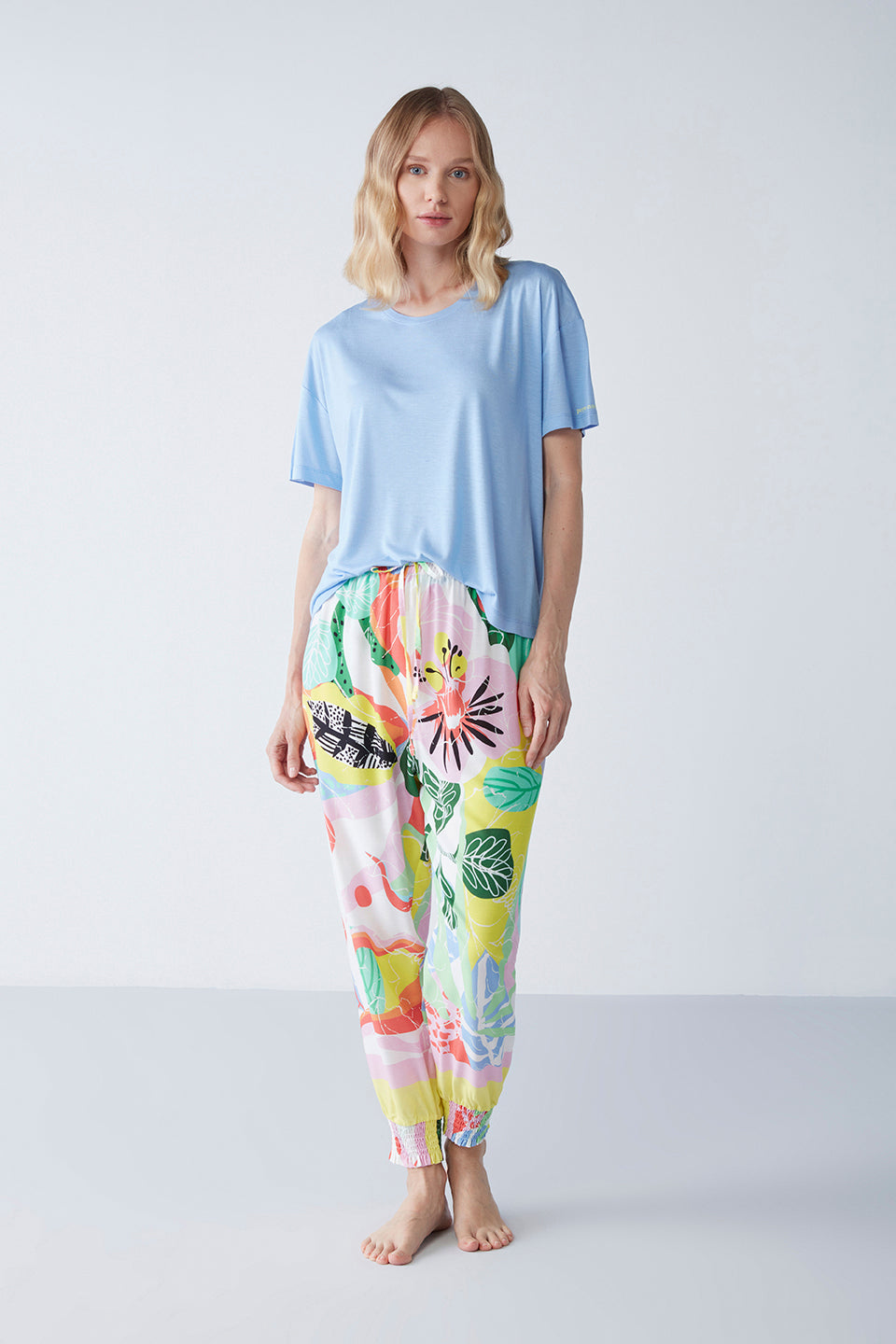 Woman wearing Popjammies "Vivid Dreams" Pyjama Set featuring a pastel blue top and vibrant, multi-colored patterned pants, standing against a white background.