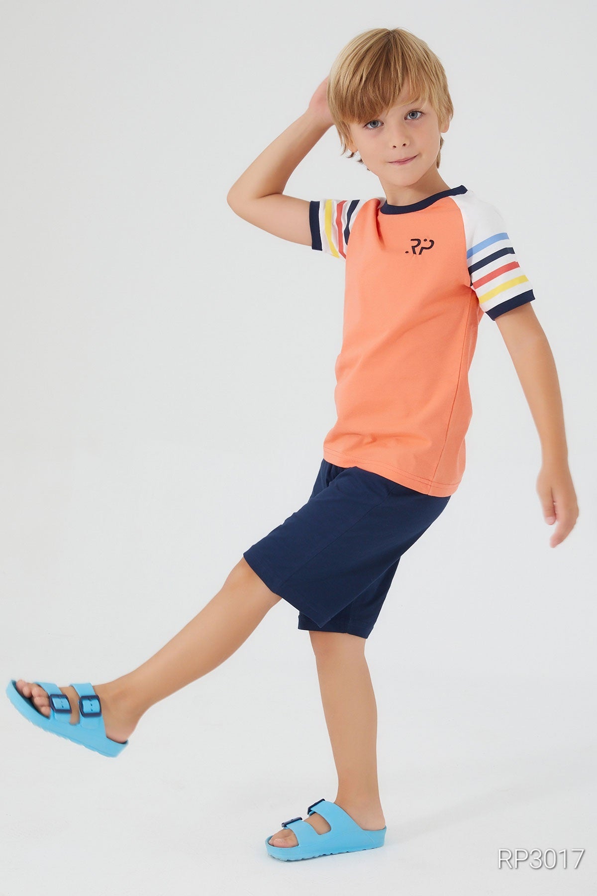  A boy posing, wearing an orange top with multicolored patterned sleeves and navy shorts, front view.