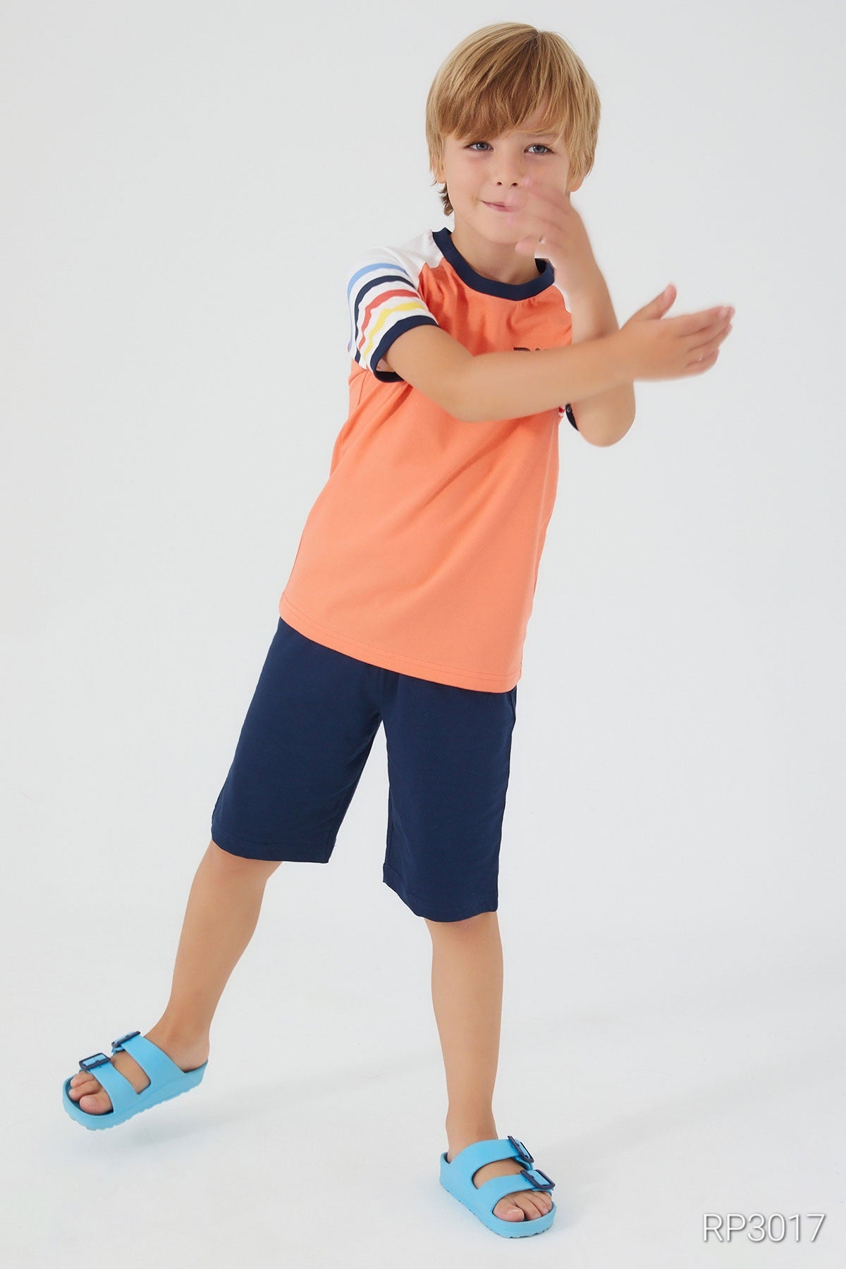 A boy with closed hands, wearing an orange top with multicolored patterned sleeves and navy shorts, front view.