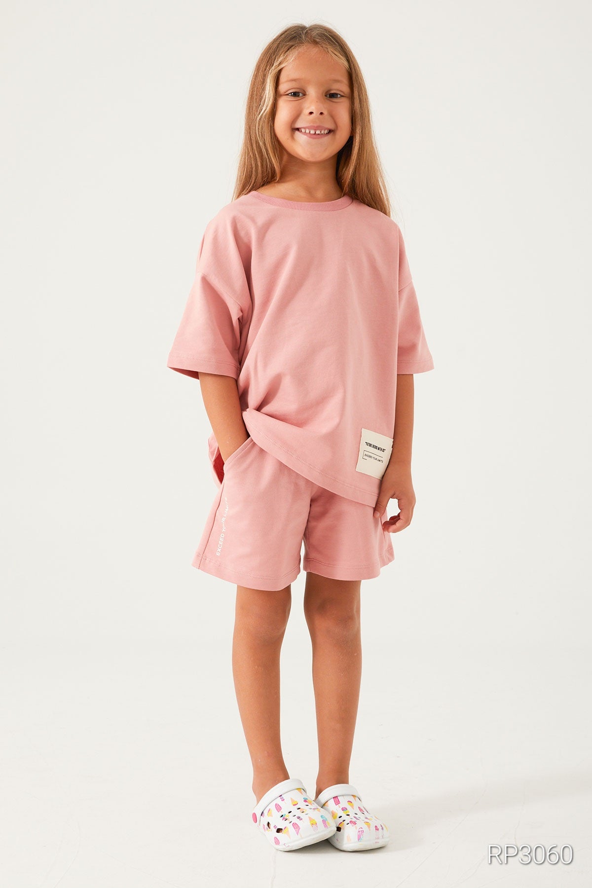 Girl smiling and wearing Popjammies Cozy Cotton Light Pink Matching Set for Girls with short sleeves and shorts, showcasing the relaxed fit and charming light pink design, with hand in her pocket