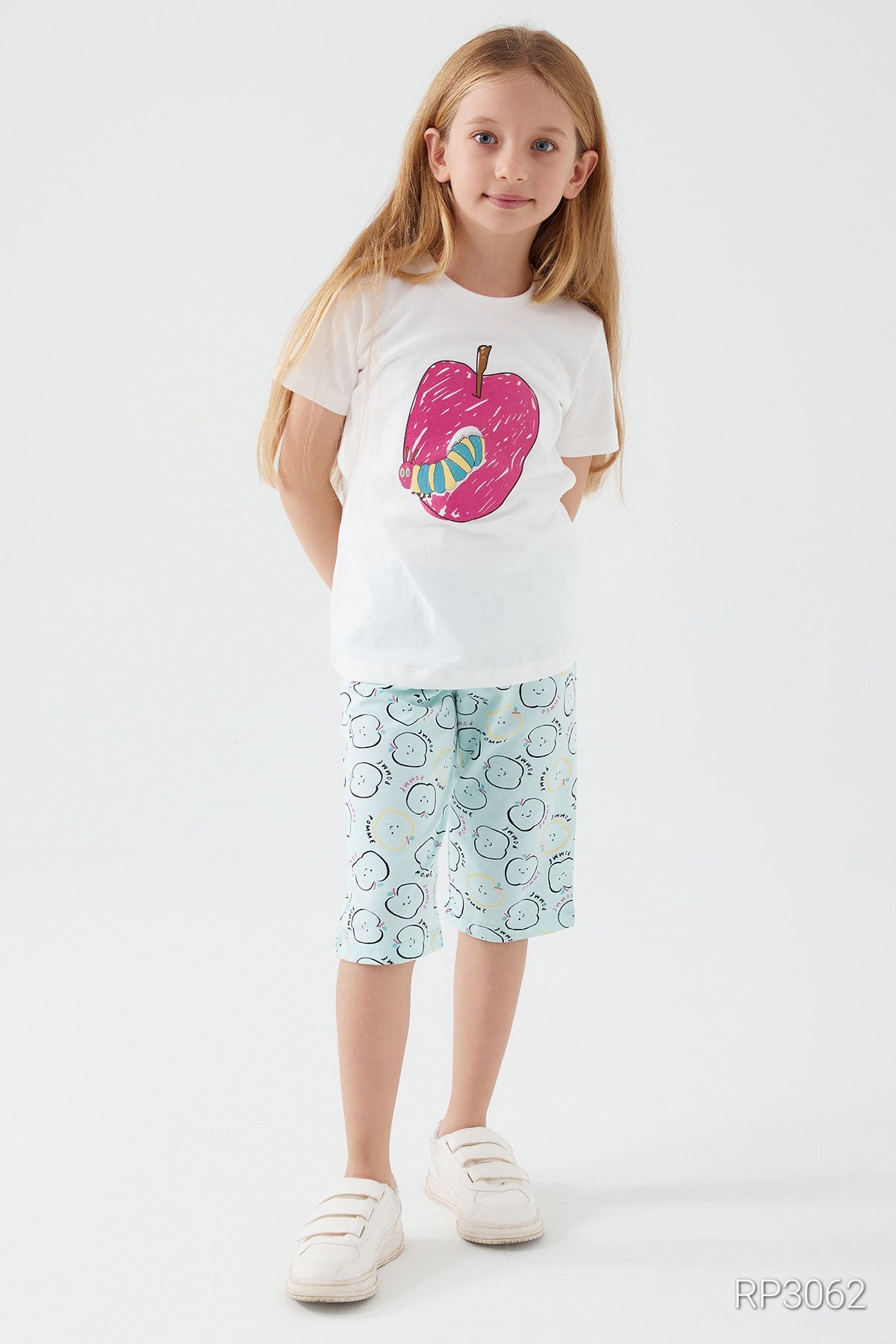 Girl wearing Popjammies Apple Delight Pajama Set with white top and light blue pantacour with small apples, with hands behind her back