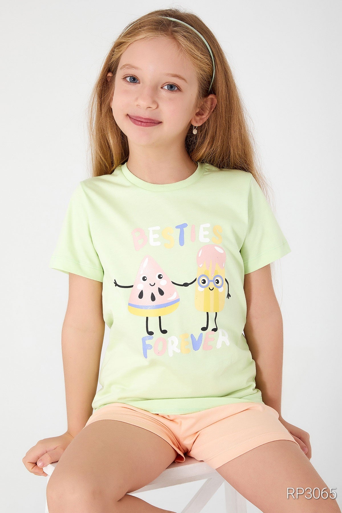 Girl sitting on a white chair wearing Popjammies 'Friends Forever' Pajama Set featuring a light green top with 'Friends Forever' text and ice cream graphic, paired with pastel-colored shorts.