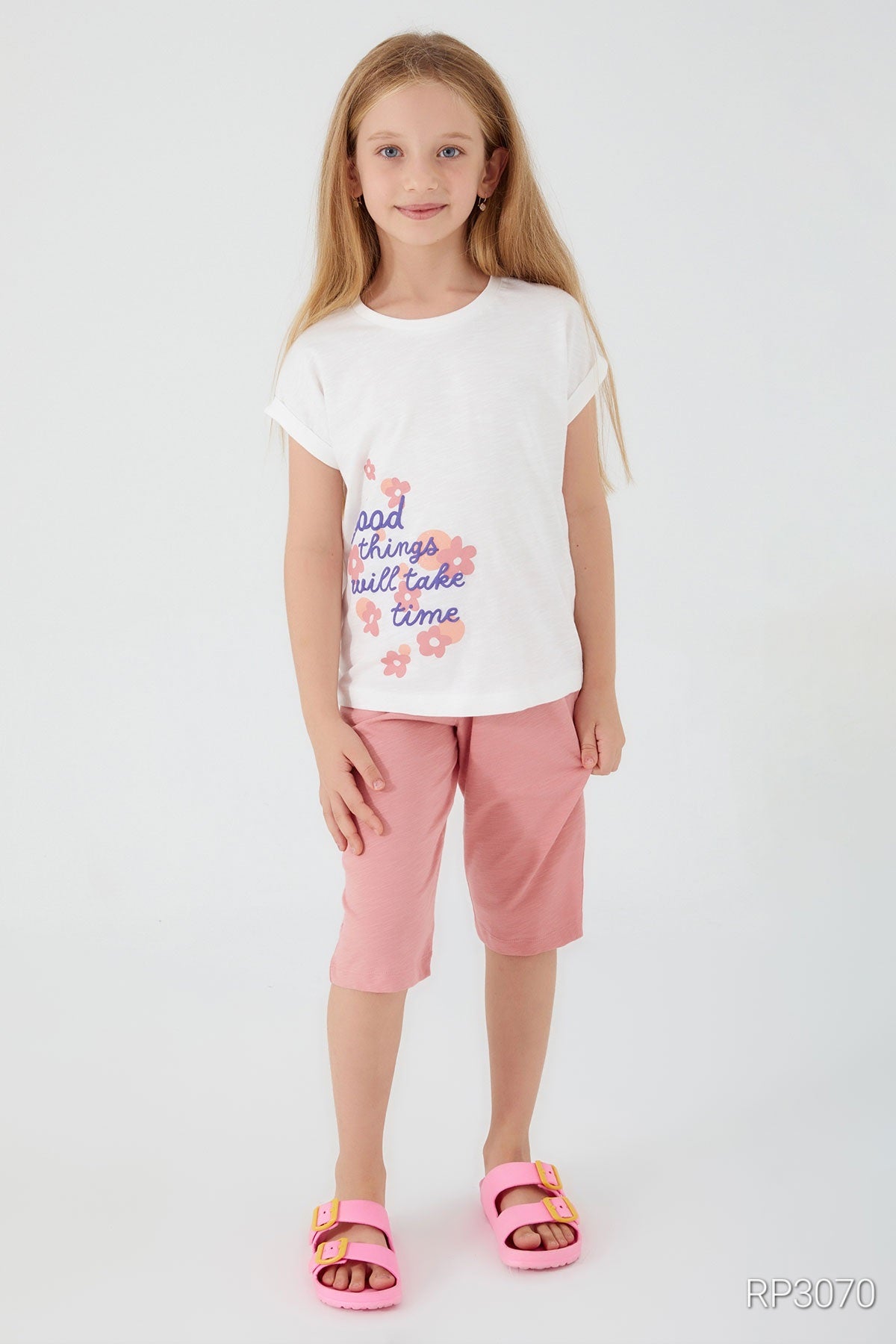 A girl wearing Popjammies 'Good Things Take Time' Pajama Set featuring a white top with 'Good Things Take Time' text and floral accents on the side, paired with light pink pantacour pants.