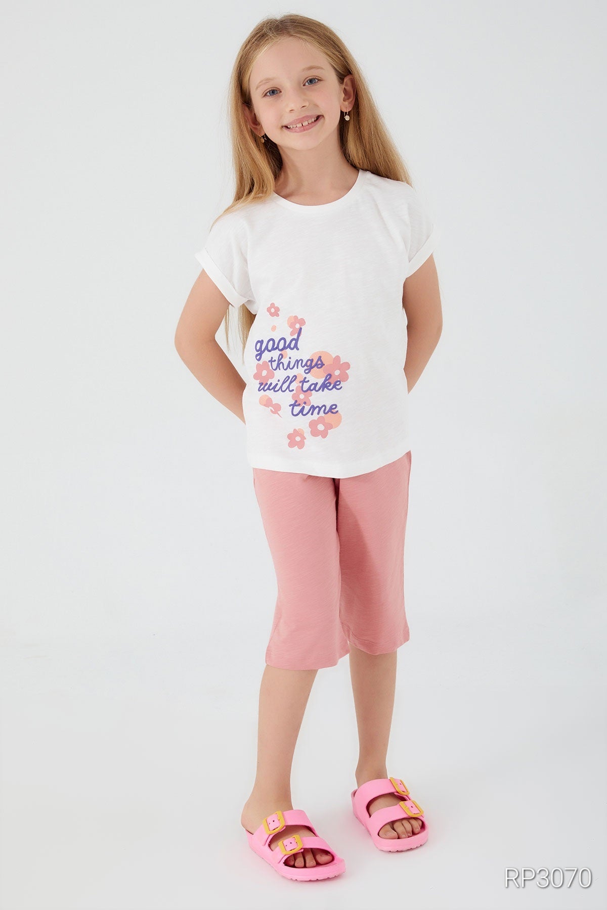 A girl wearing Popjammies 'Good Things Take Time' Pajama Set featuring a white top with 'Good Things Take Time' text and floral accents on the side, paired with light pink pantacour pants, smiling