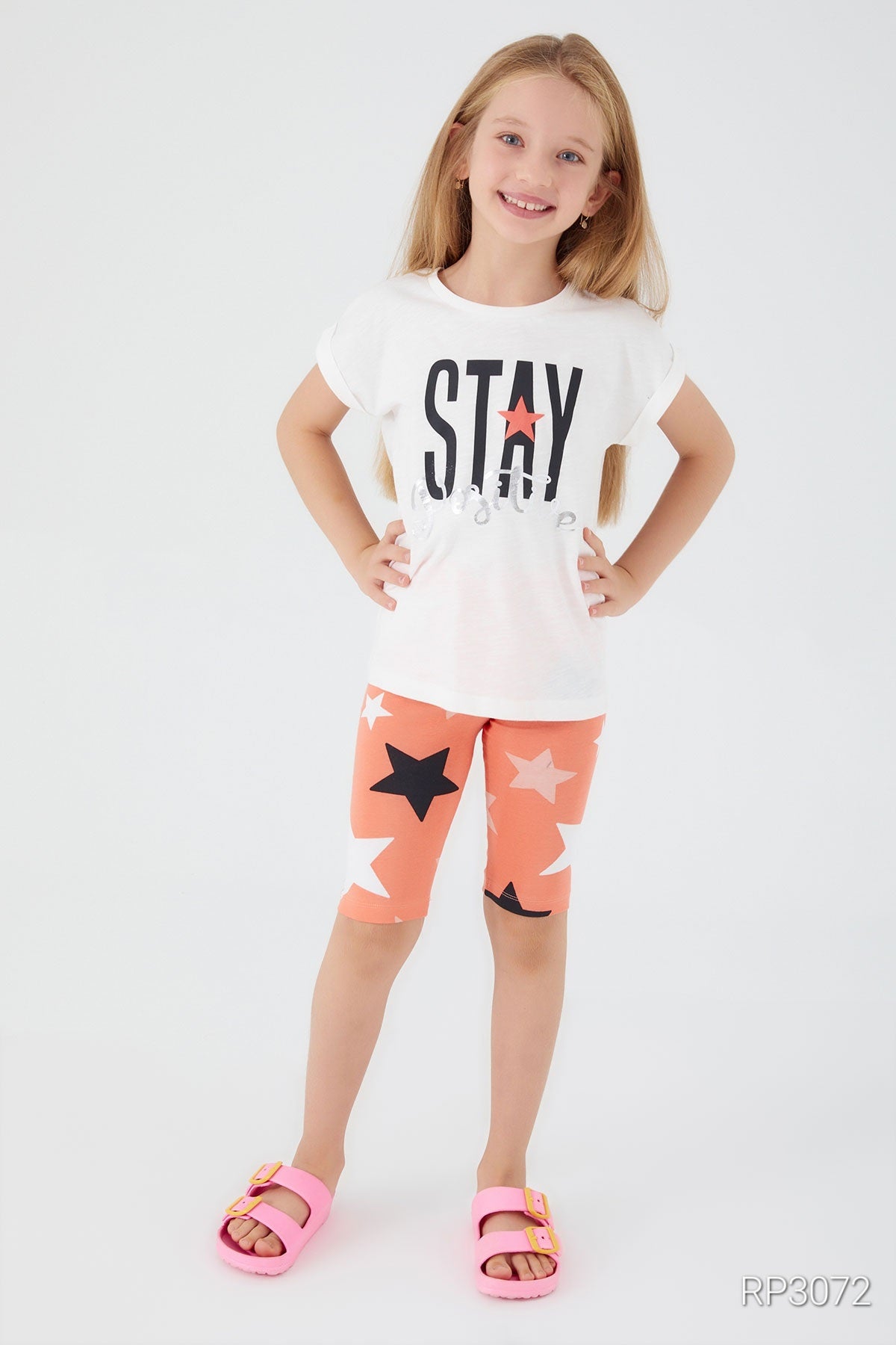 Front view of Popjammies 'Stay Positive' Pajama Set featuring a white top with 'Stay Positive' text and orange legging shorts with stars, smiling with hands on her hips