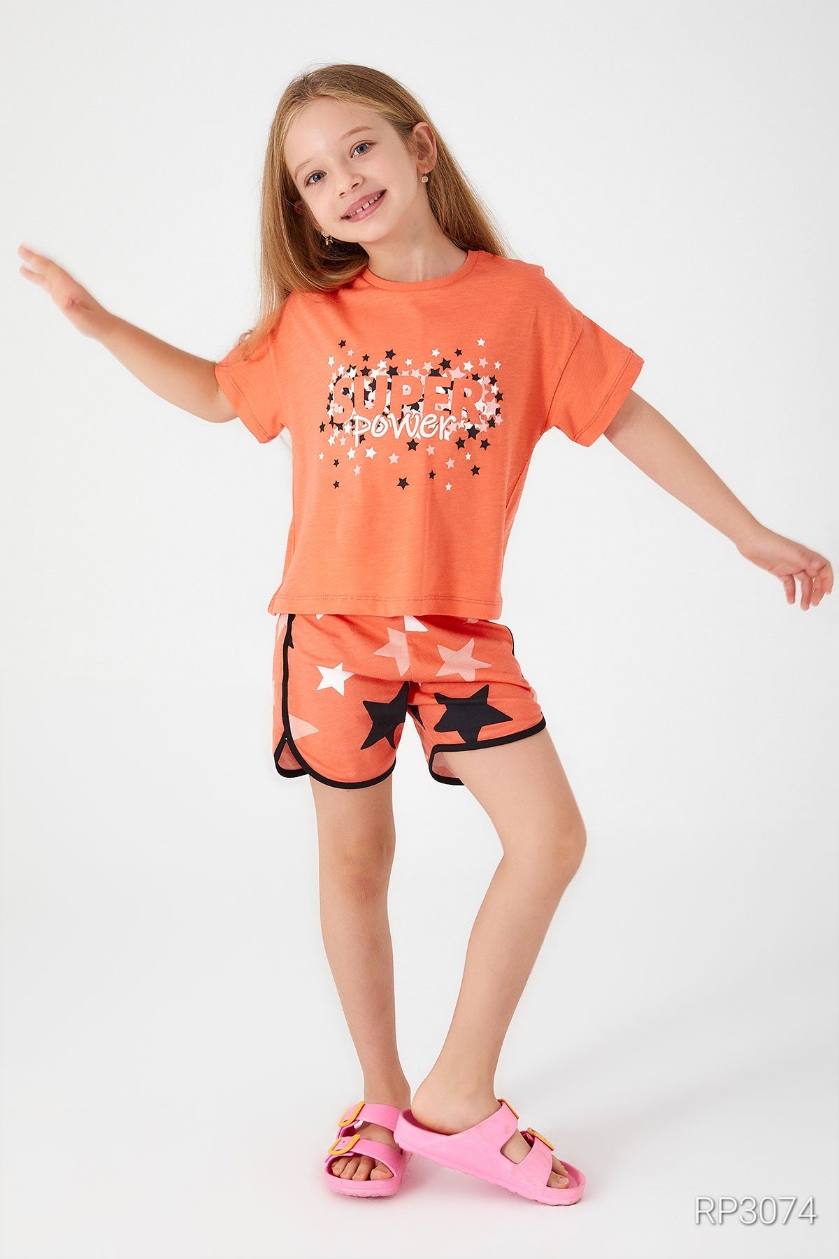 Girl wearing an orange top with "Super Girl" written on it and matching orange shorts with star patterns in front of a plain background.