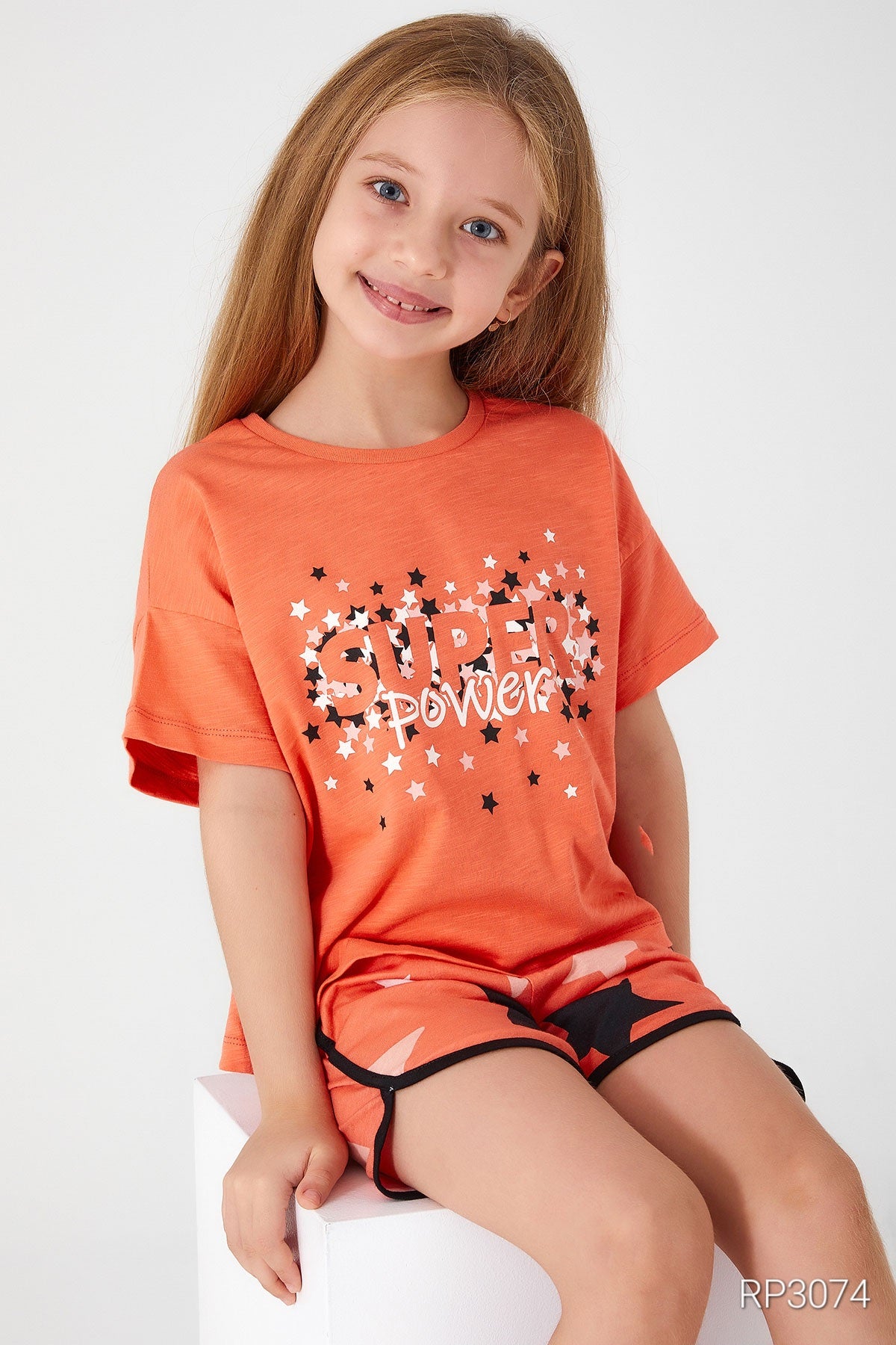 Girl wearing an orange top with "Super Girl" written on it and matching orange shorts sitting on a white shorts.