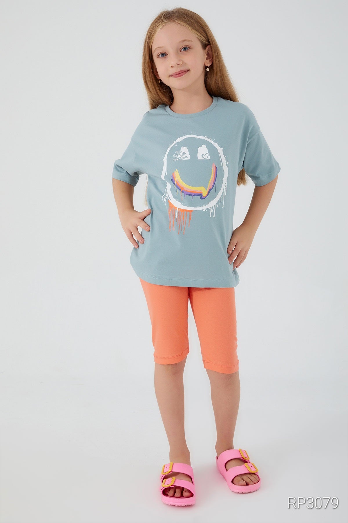 Girl wearing a baby blue top with a smiley face and orange short leggings, with hands on hips