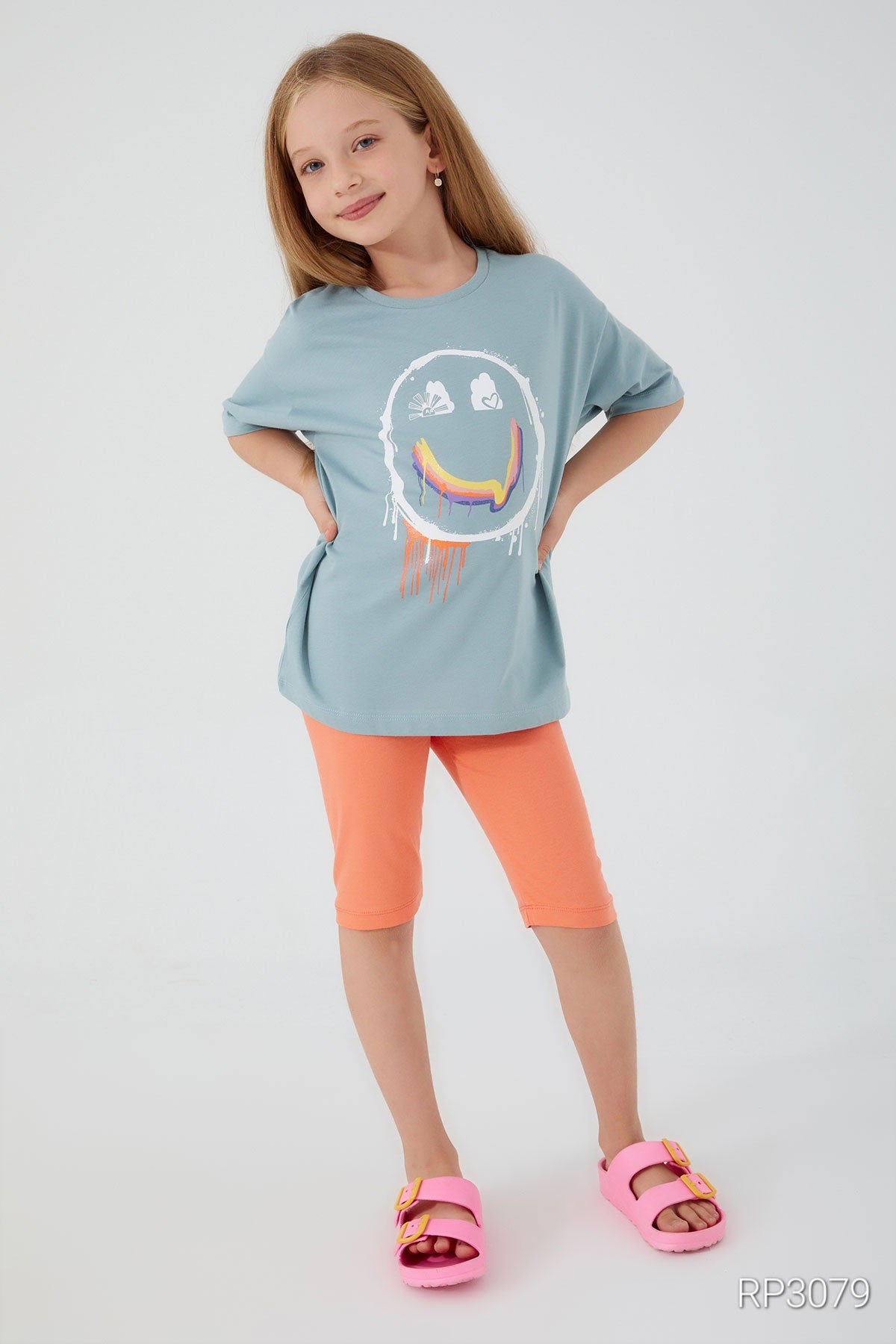 Girl wearing a baby blue top with a smiley face and orange short leggings, smiling with hands on her hips.