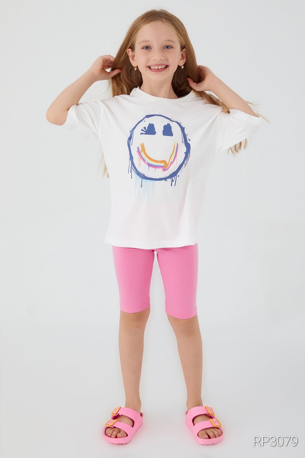 Girl holding her hair and wearing a white short sleeve top with a smiley face and pink legging shorts in front of a white background