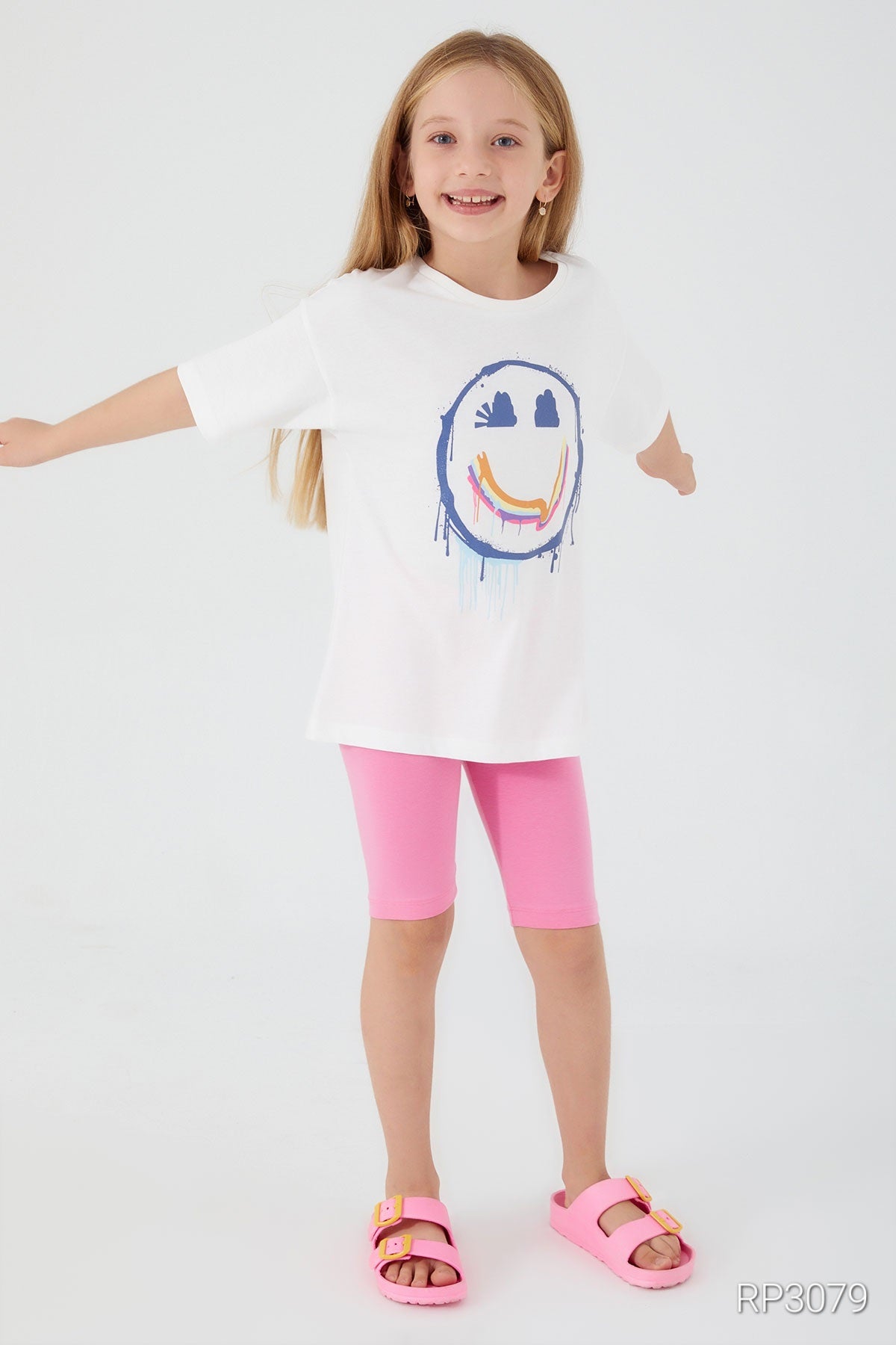 Girl wearing a white short sleeve top with a smiley face and pink legging shorts in front of a plain background