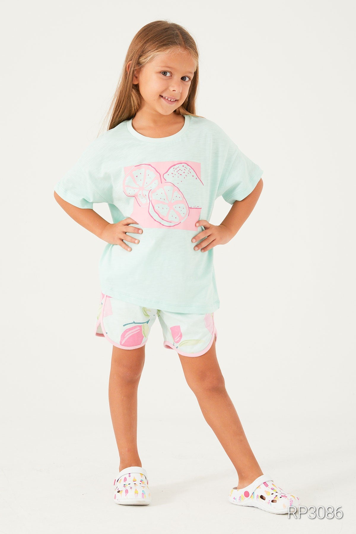 Girl wearing Popjammies Lemon Fresh Pajama Set with baby blue top and white shorts with pink lemons, smiling with hands on hips