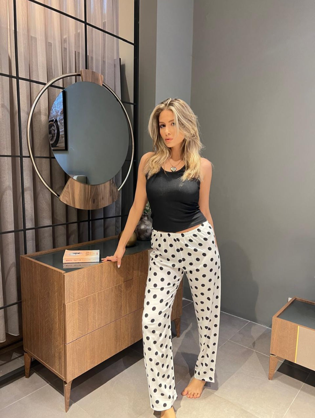 Model wearing a black tank top and white pyjama pants with black polka dots from Popjammies, standing with one hand resting on a wooden dresser in a modern room with a circular mirror and decorative elements.